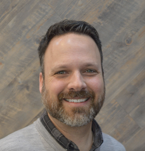 A smiling man with a beard and short hair, wearing a gray sweater over a plaid shirt, standing against a wooden background.