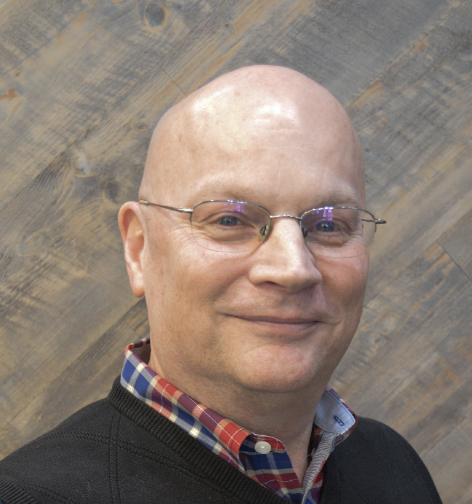 A bald man wearing glasses, a black sweater, and a plaid shirt, smiling in front of a wooden wall background.