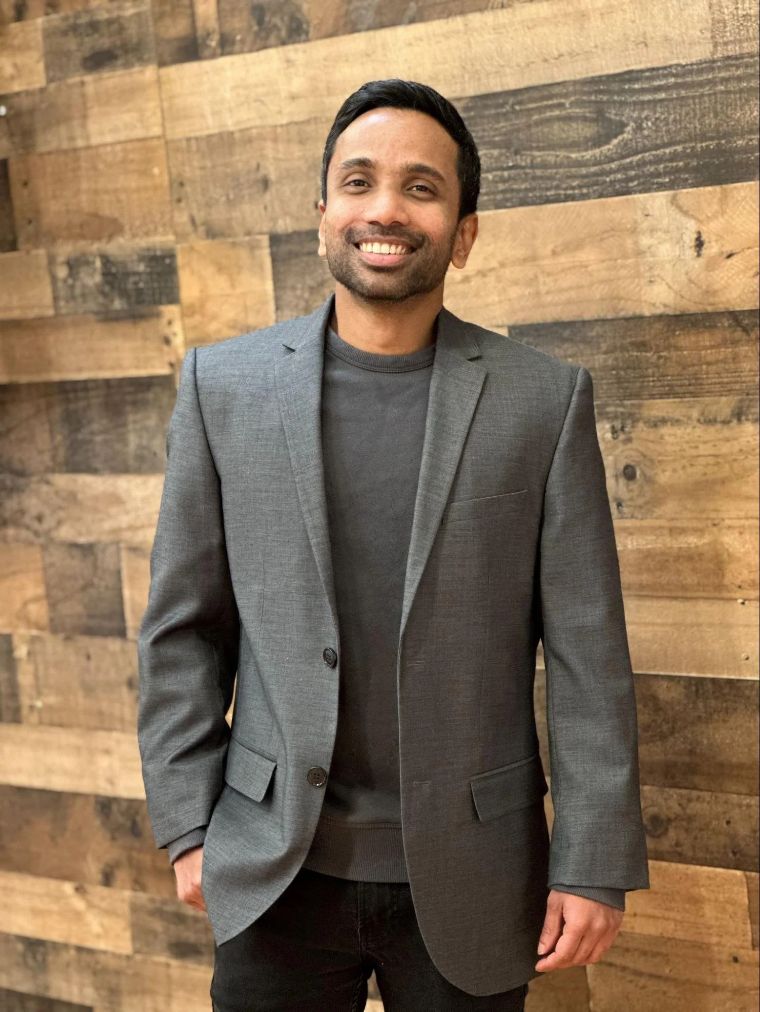 A smiling man in a gray blazer and black shirt standing against a wooden wall with a mix of light and dark wood planks.