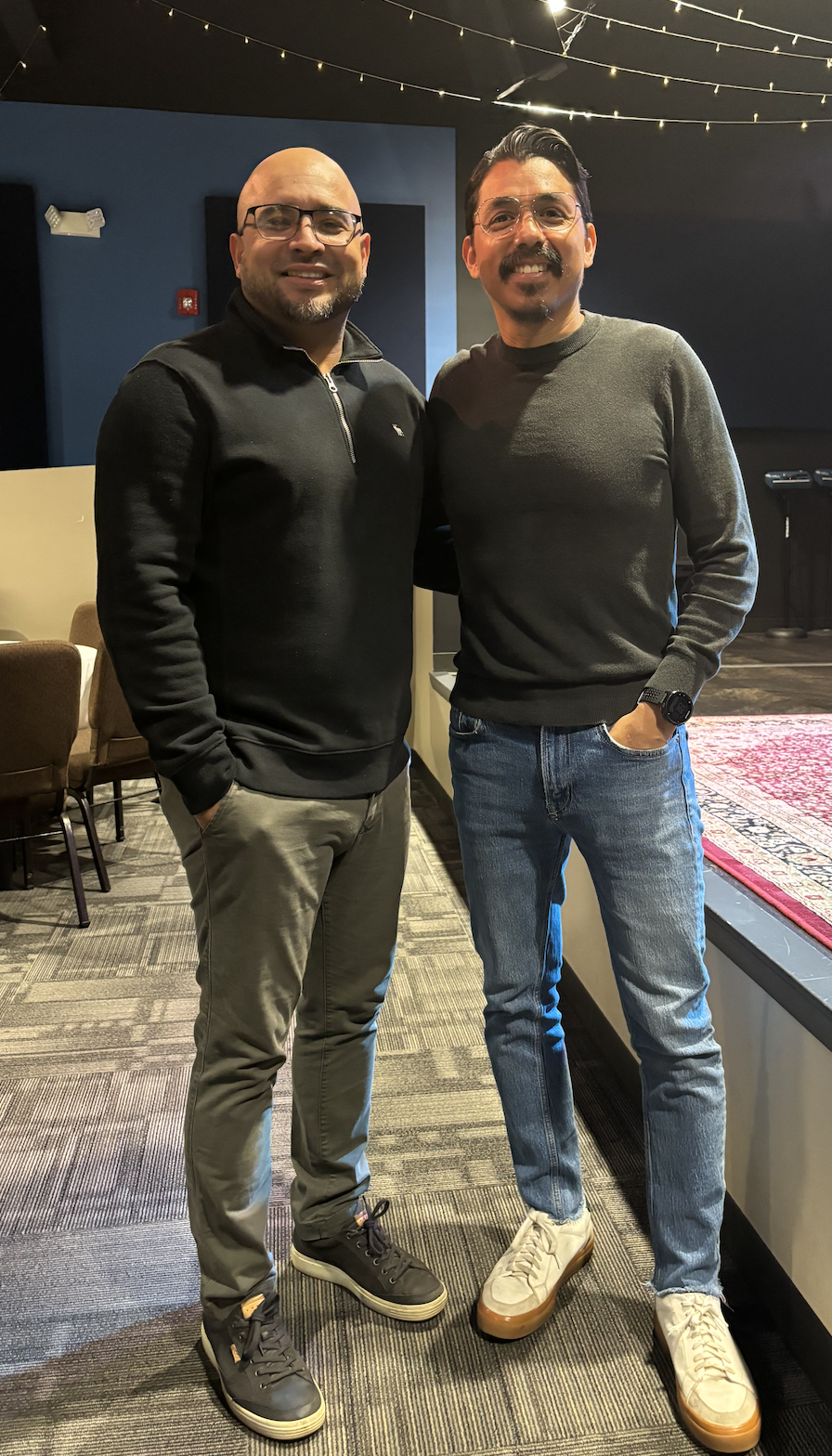 Two men standing close together, smiling at the camera, in an indoor setting with string lights overhead and chairs and a stage area in the background.