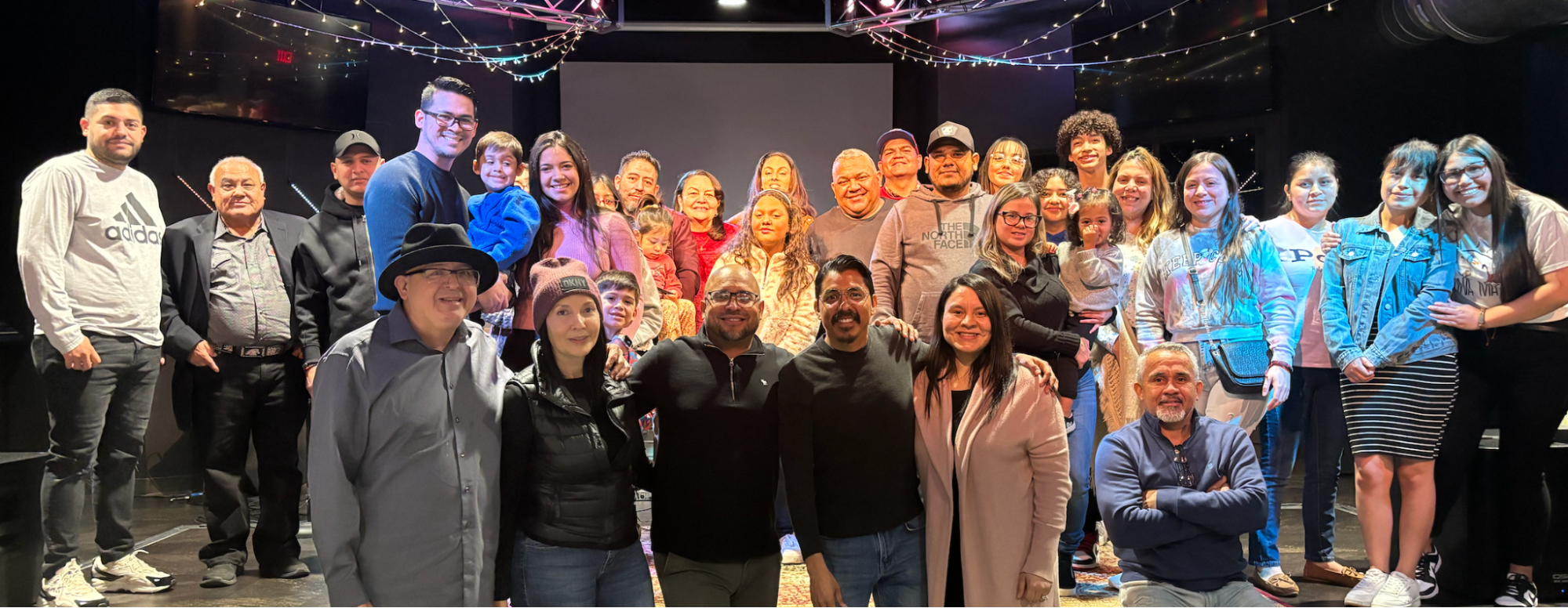 Group of people of various ages and backgrounds, gathered on stage in a theater or auditorium, smiling for a group photo. Ceiling decorated with string lights, background shows a large blank screen.