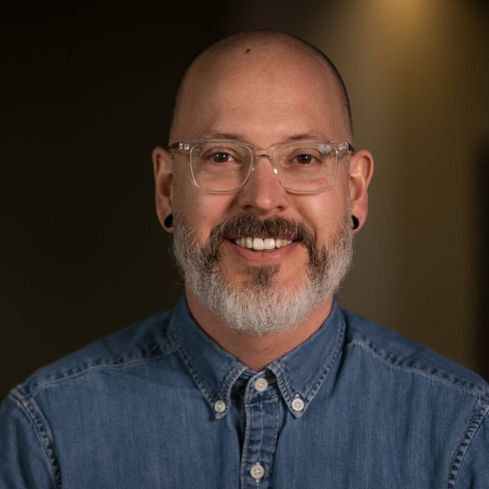 A smiling man with glasses, a beard, and earrings, wearing a denim shirt.