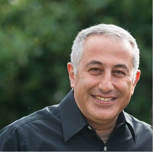 A smiling middle-aged man with short gray hair wearing a black button-up shirt outdoors.
