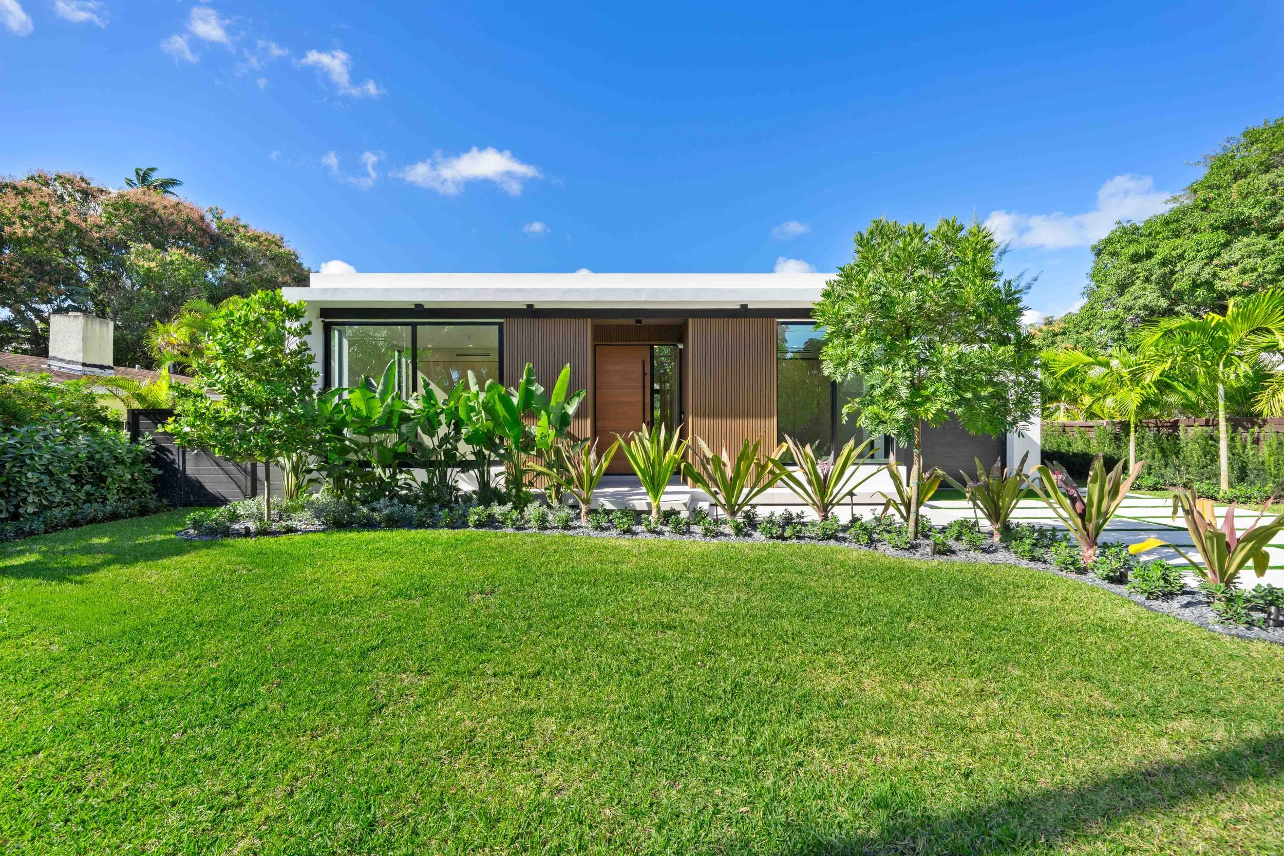 Modern white house with large glass windows, surrounded by green trees and a neatly manicured lawn under a blue sky.
