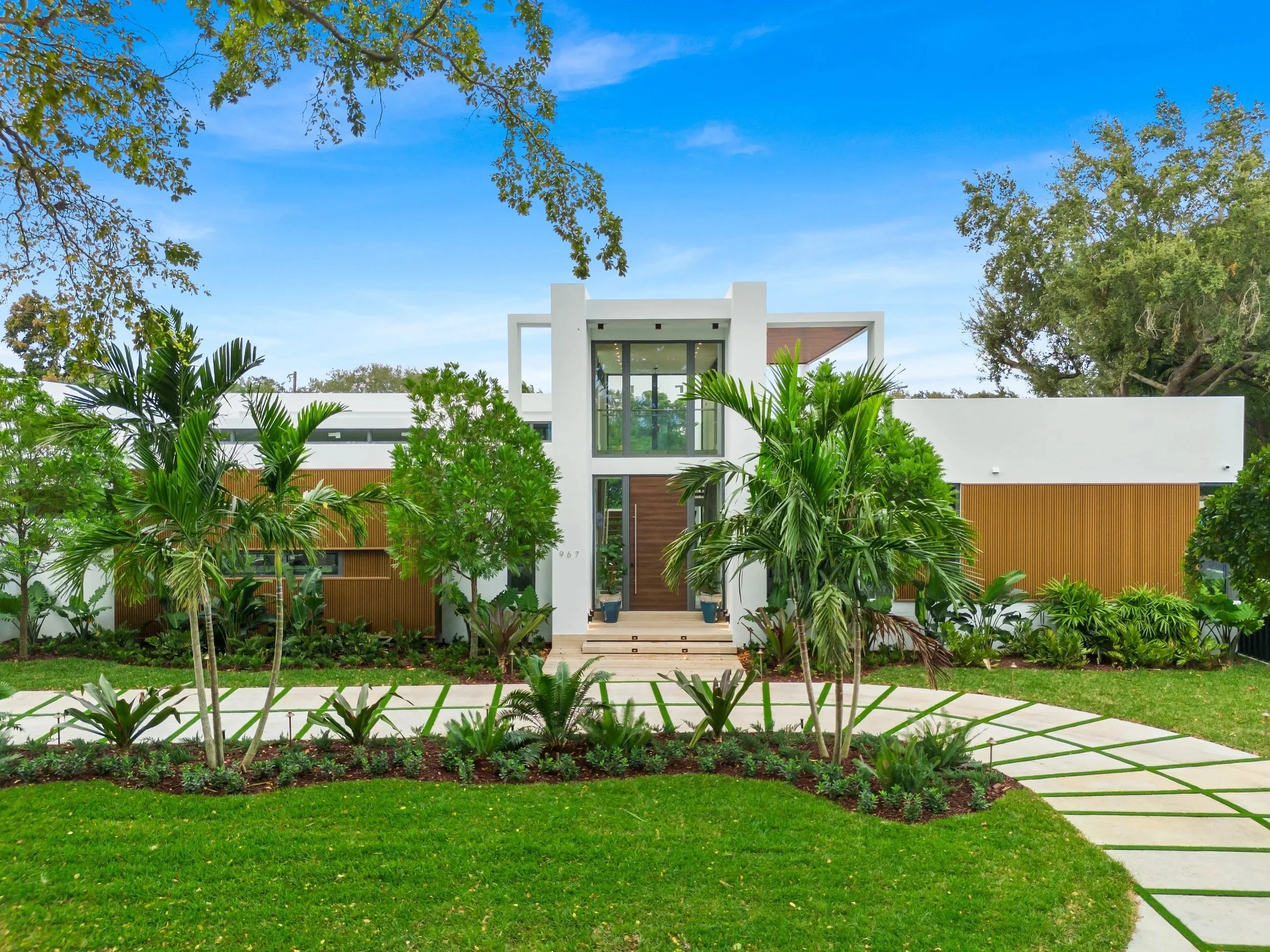 Modern house with white exterior, wooden accents, and large glass windows, surrounded by lush greenery and a curved stone pathway.