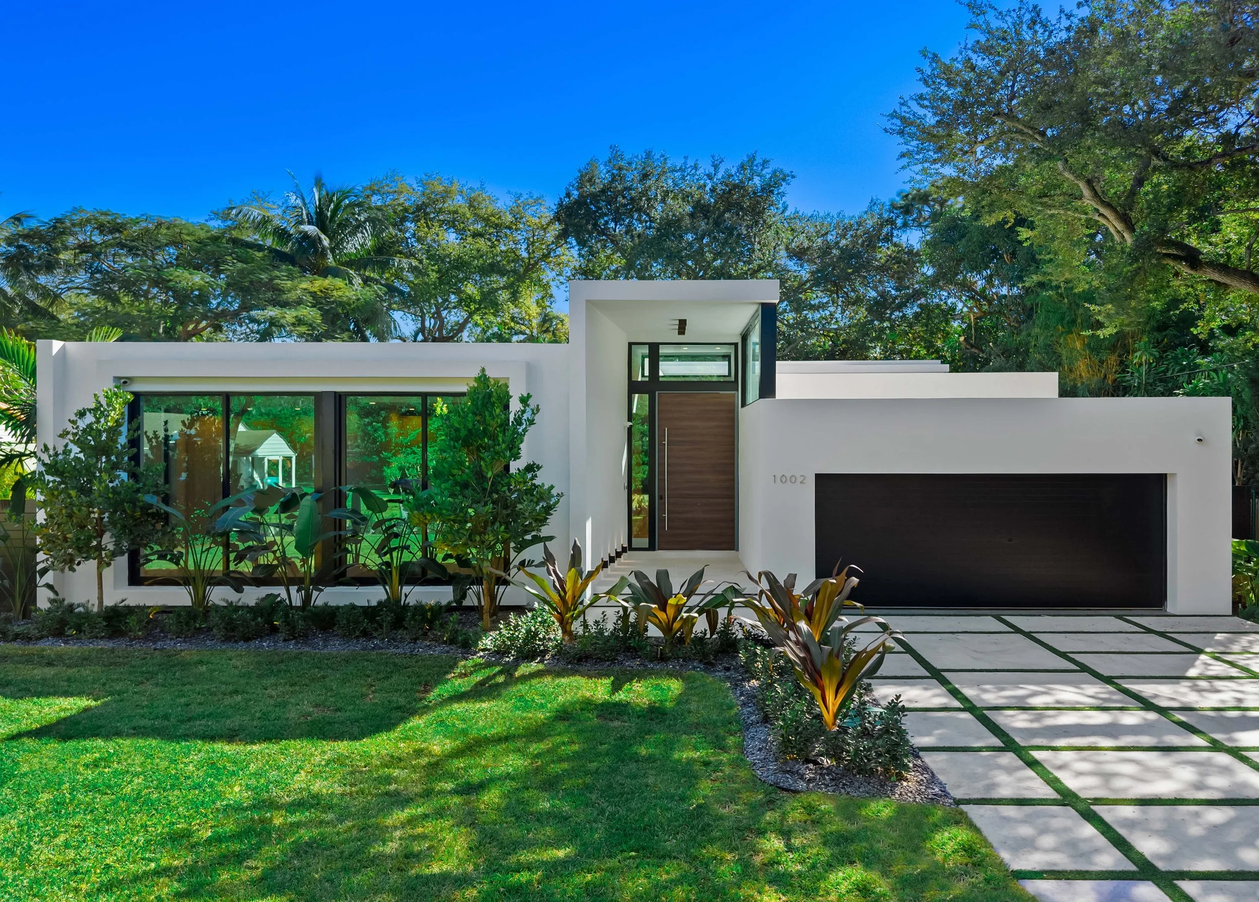 Modern white house with large glass windows, a dark garage door, and a front yard with green grass, small bushes, and a tiled pathway, under a clear blue sky.