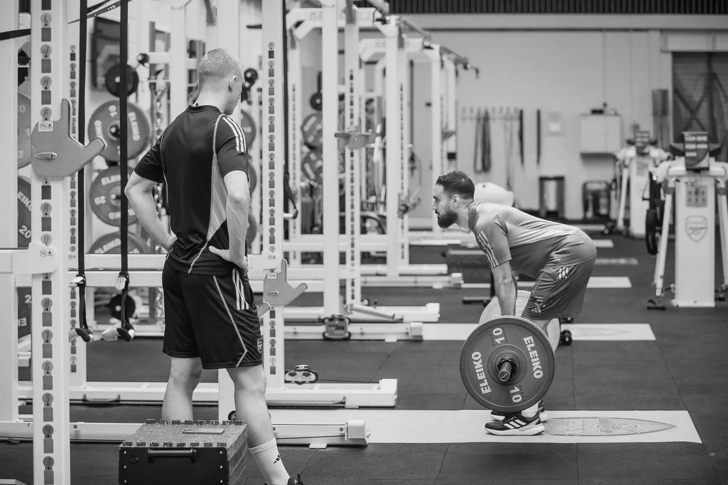 Two men in a gym, one standing with hands on hips, another bending down lifting a barbell, surrounded by gym equipment.