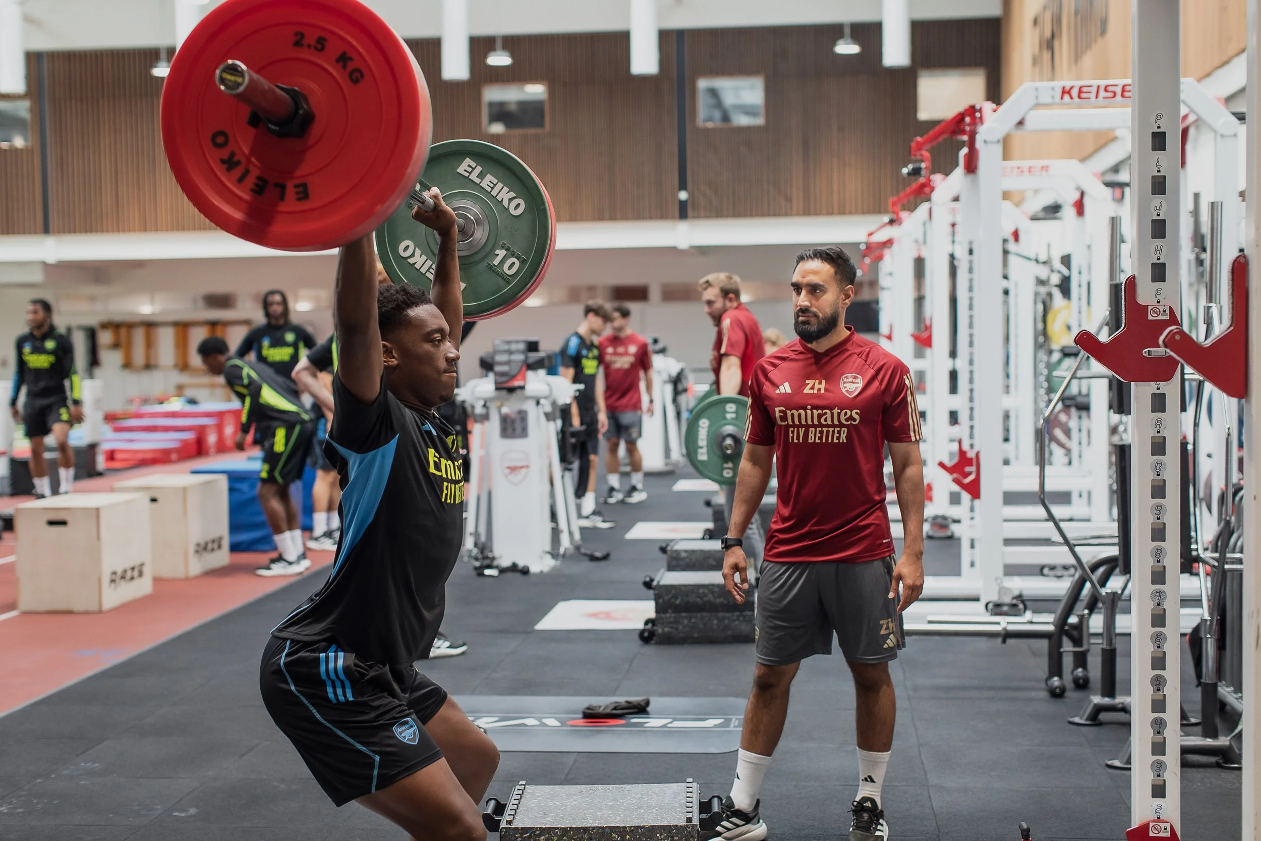 A male athlete performs a barbell overhead press in a gym, guided by a coach. The gym has various exercise equipment and other athletes working out in the background.