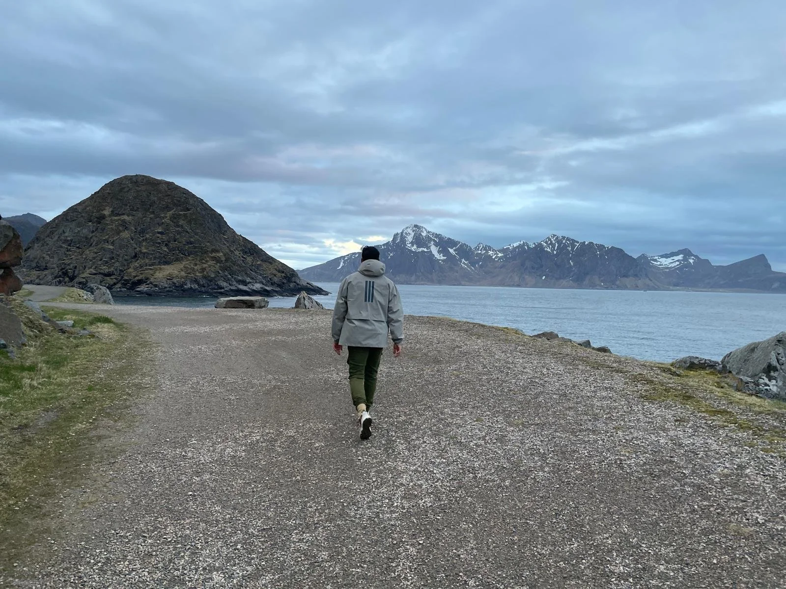 A person walking along a gravel path by a body of water with mountains in the background, under a cloudy sky.