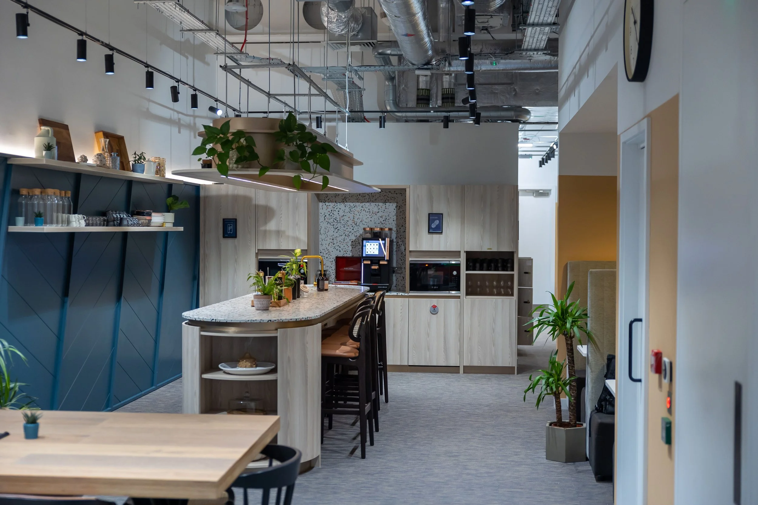 Modern office kitchen with a breakfast bar, plants, and coffee machine, featuring wood cabinets and neutral decor.