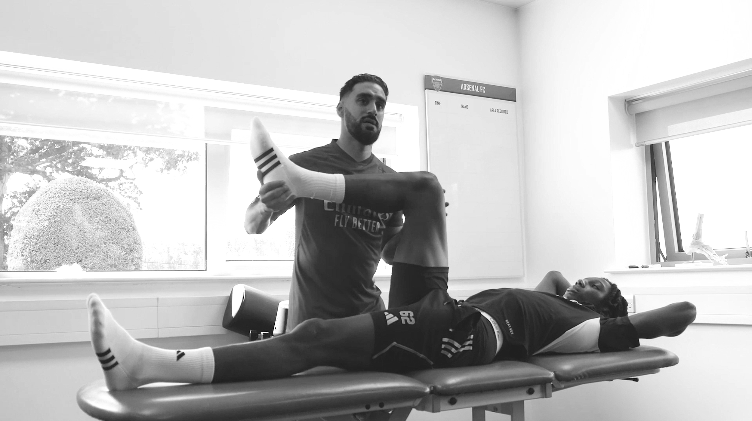 A physical therapist or trainer performing a stretching or medical assessment on a male athlete lying on an examination table, with a window showing trees outside in the background.