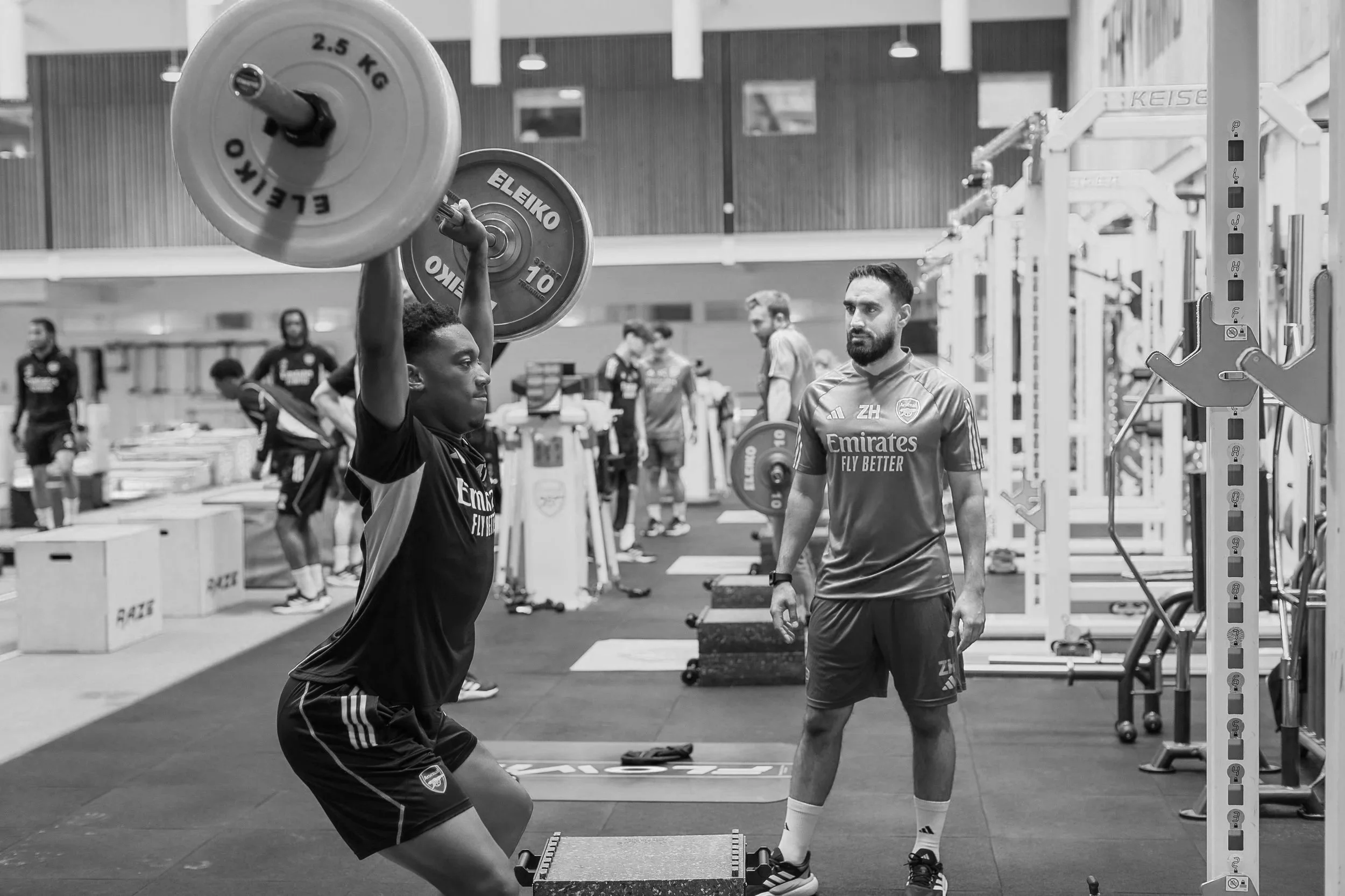 A man lifting a barbell above his head in a gym with a coach observing and other athletes working out in the background.