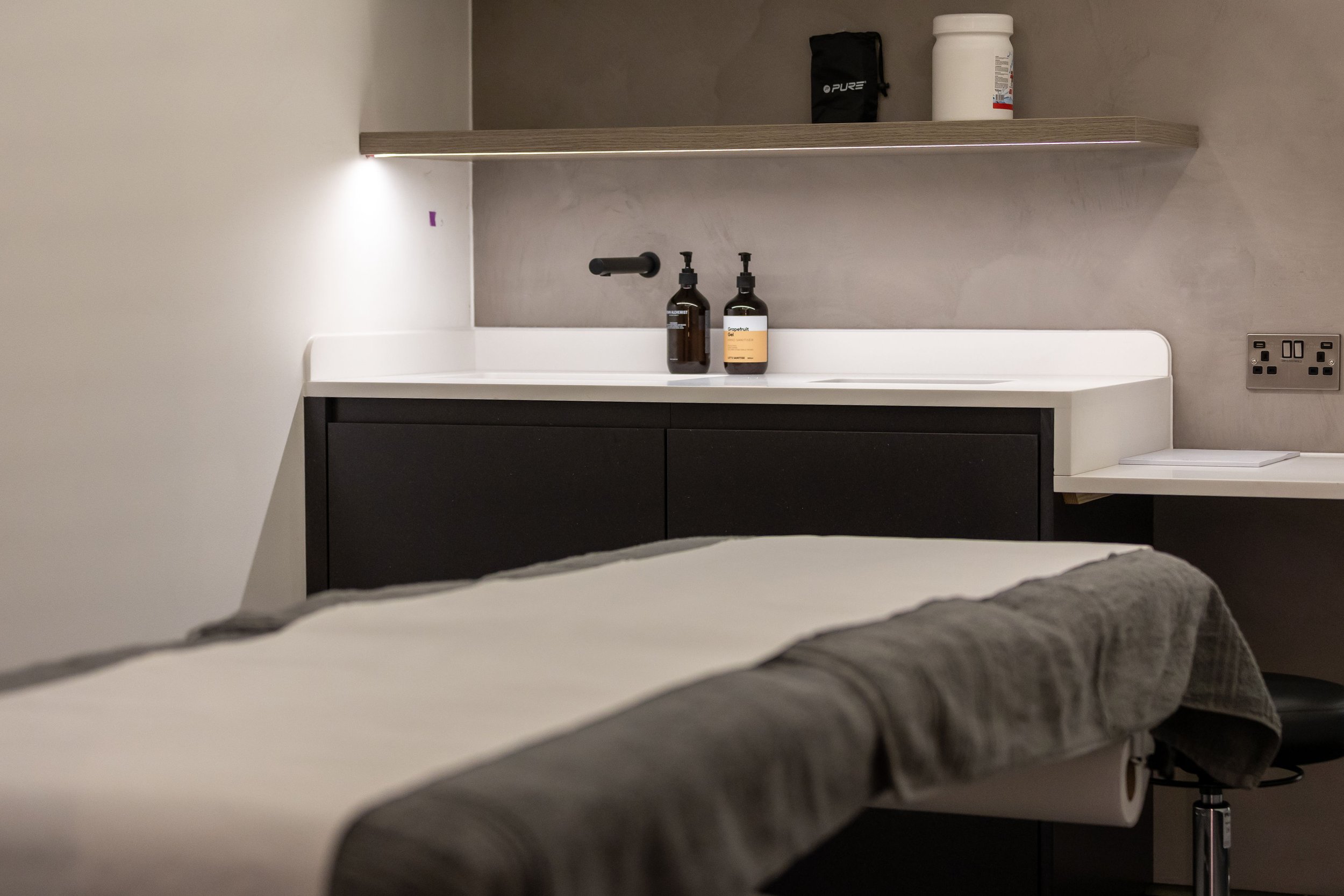 A modern medical or massage room with a bed covered in a brown towel, a white countertop with two bottles of essential oils or lotion, and a wall-mounted shelf with a black container and a white container. There are electrical outlets on the wall.
