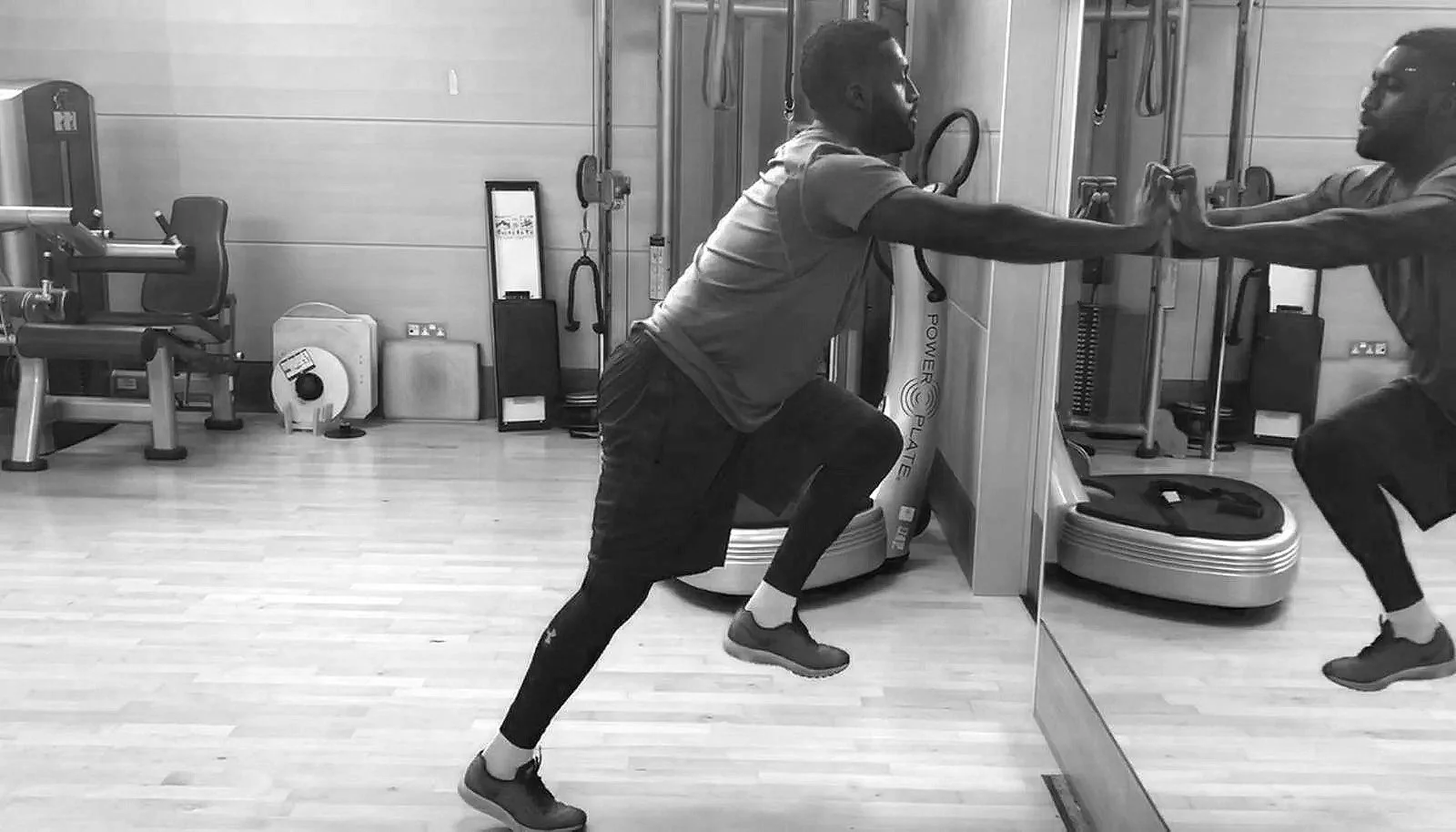 Two men exercising in a gym, one holding a plank position with one leg raised and extending arm to touch the other man’s hand, who is balancing while holding the first man's hand.