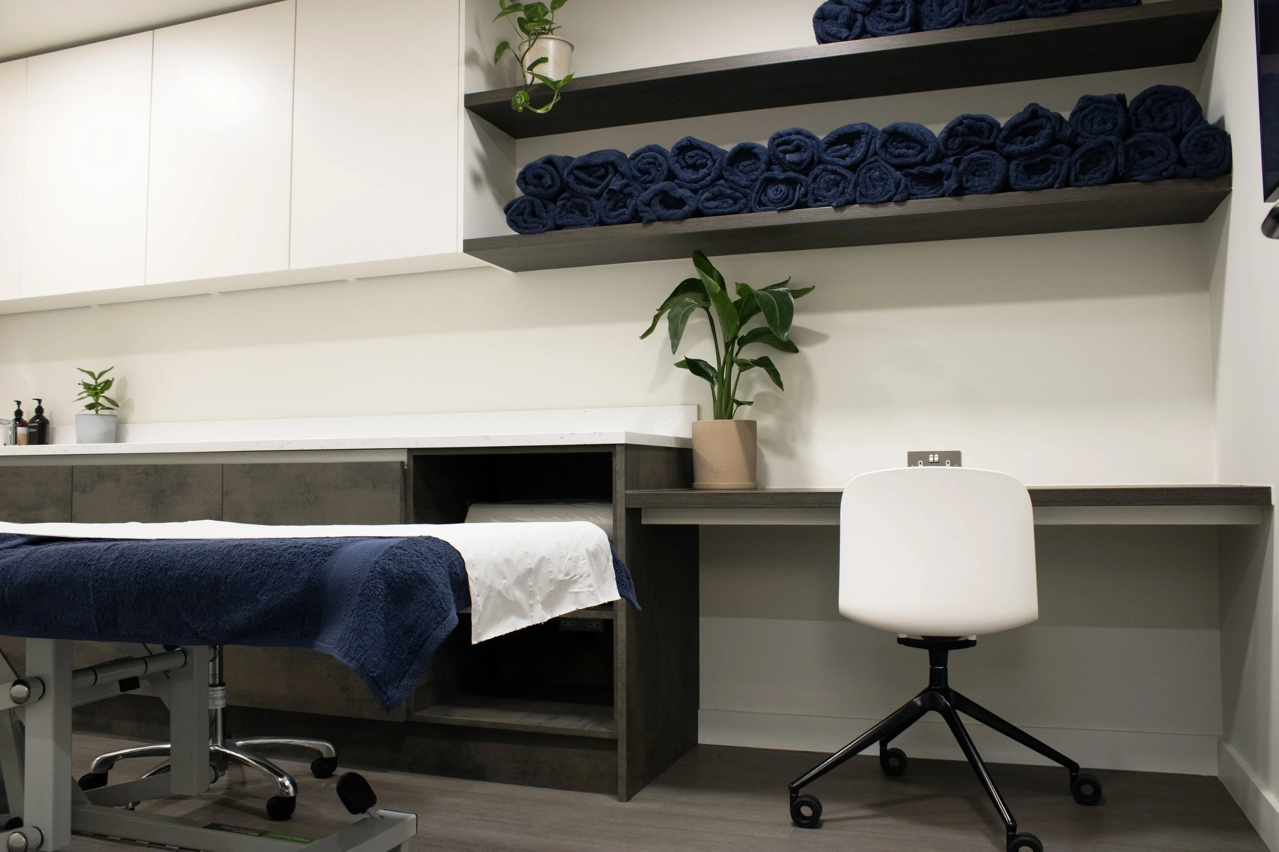 Interior of a wellness or massage room with a massage table covered in a navy towel, a white rolling chair, shelves with rolled navy towels, potted plants, and a white cabinet.