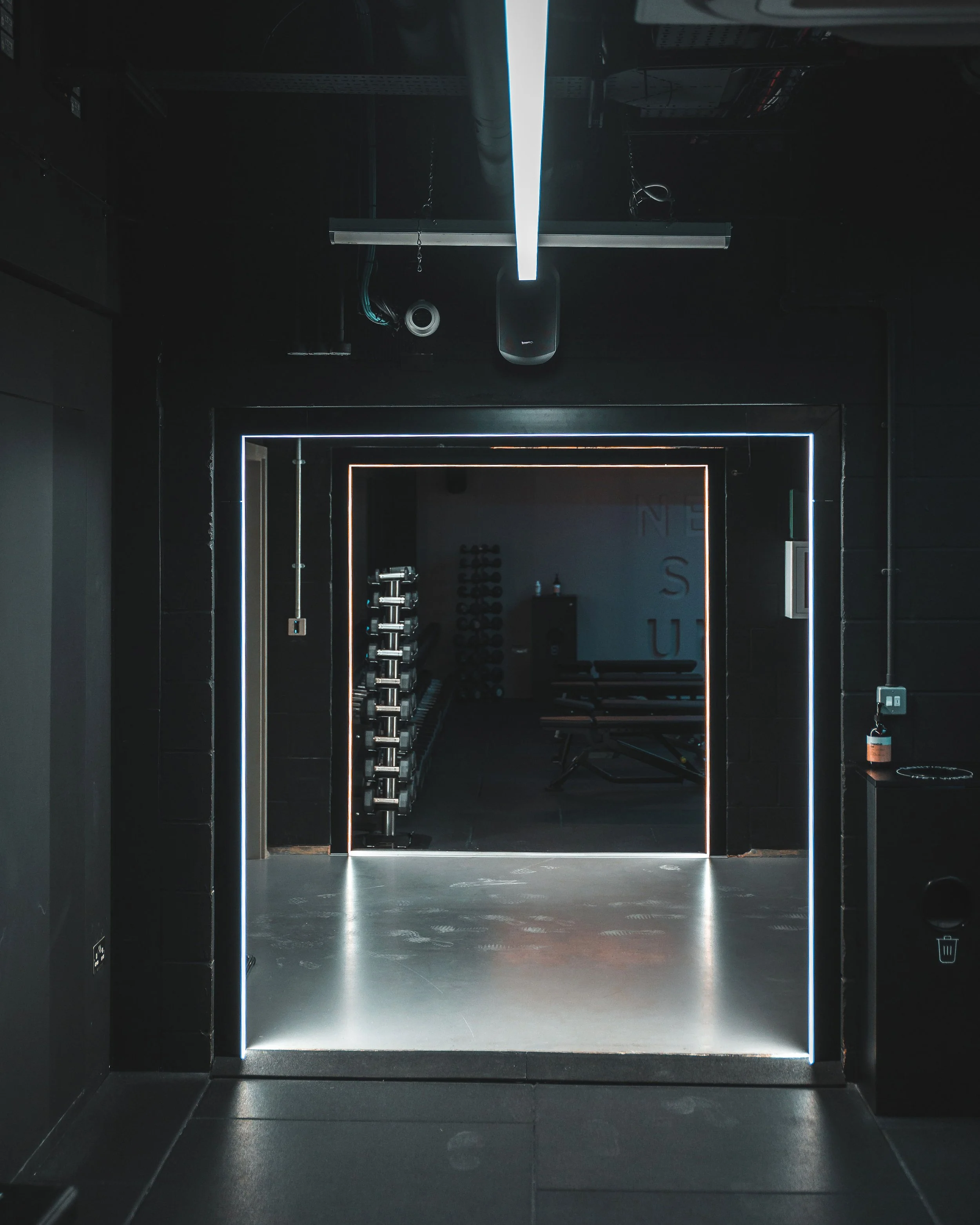 A dark gym entrance with illuminated door outlines, leading to a room with dumbbells and workout equipment visible inside.