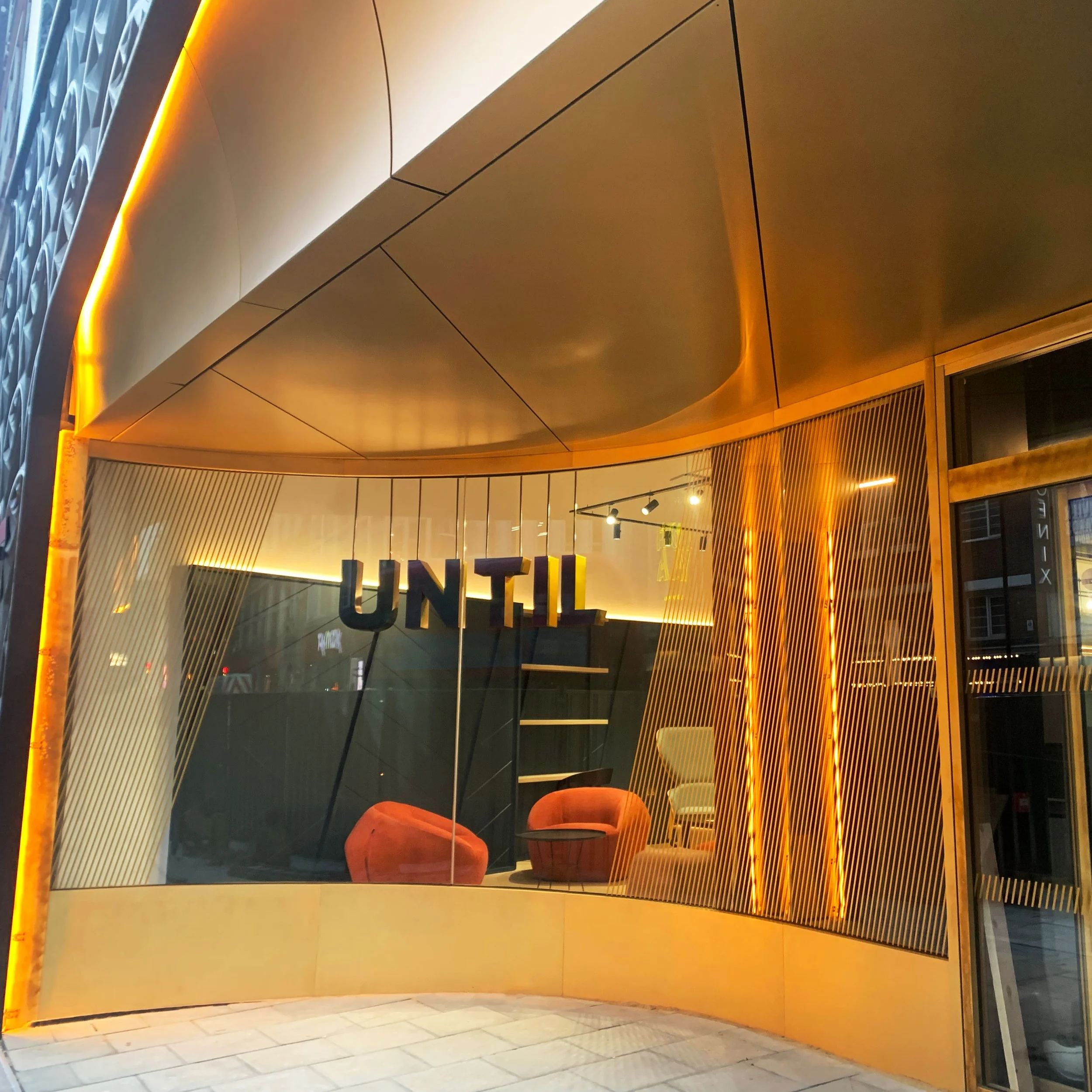 Modern storefront with orange and gold interior design, featuring large glass windows, orange chairs, and a sign that reads 'UNTIL' inside.