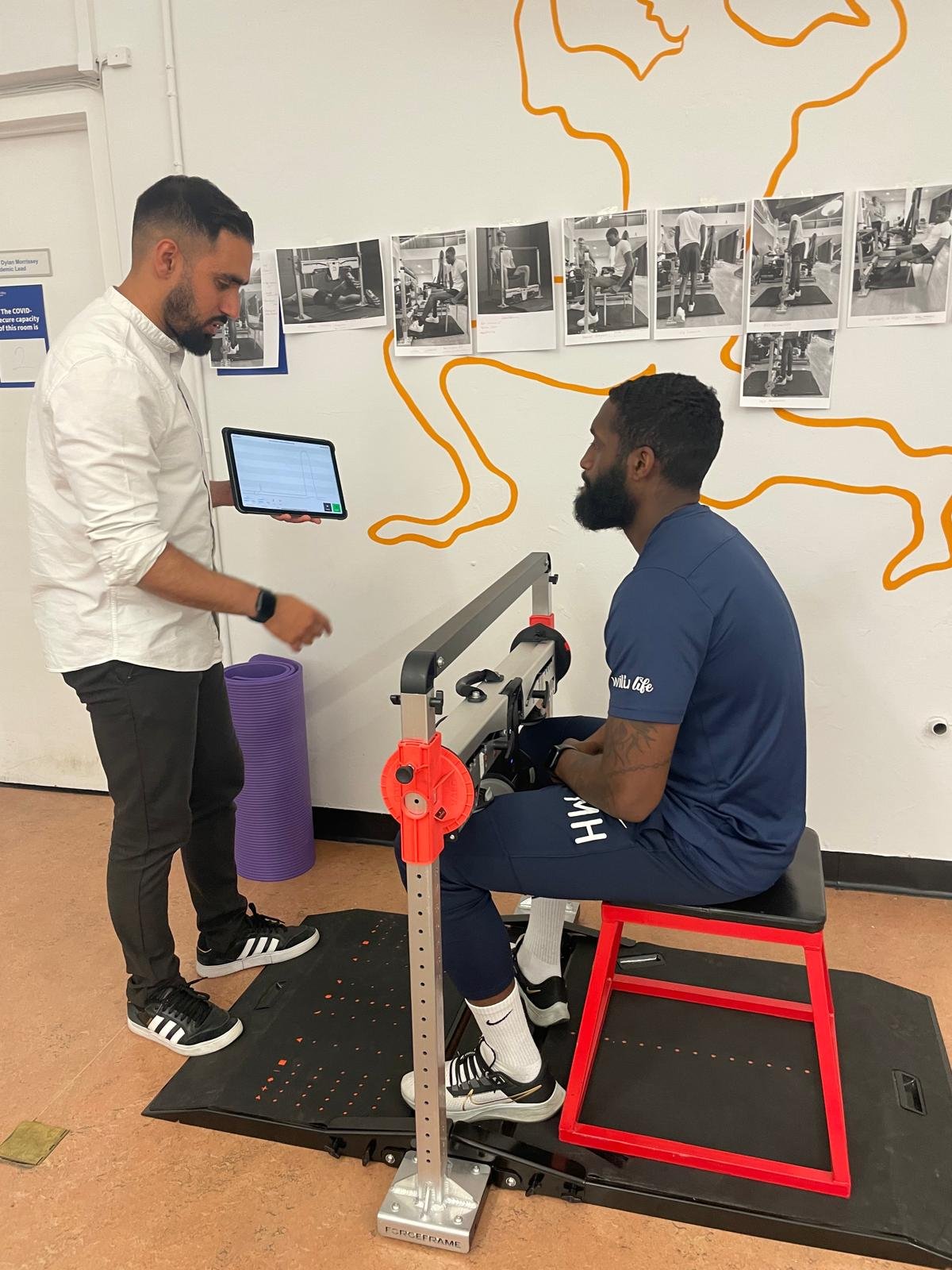 A man sitting on a red bench performing a physical test using a specialized device while another man stands nearby holding a tablet. Photographs of exercise routines are posted on the wall behind them.
