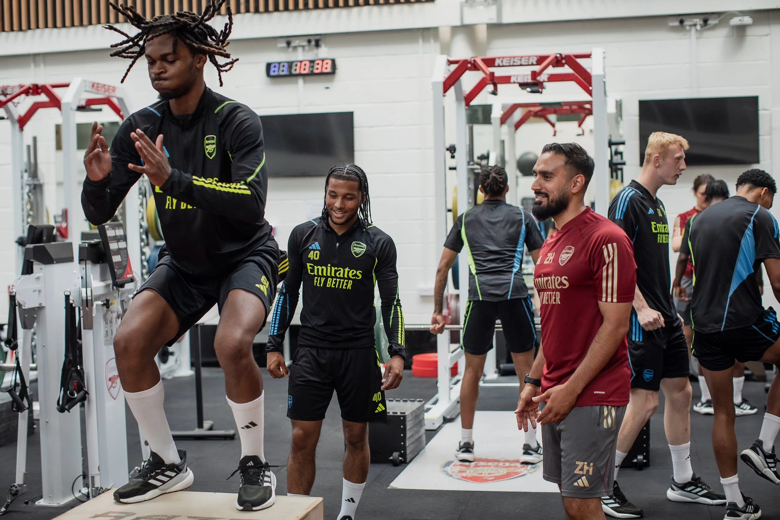 A group of soccer players training in a gym. One player is mid-jump onto a box, wearing black training gear. Others are watching, smiling, or engaging in workout activities. The gym has fitness equipment, a digital clock, and a white brick wall.