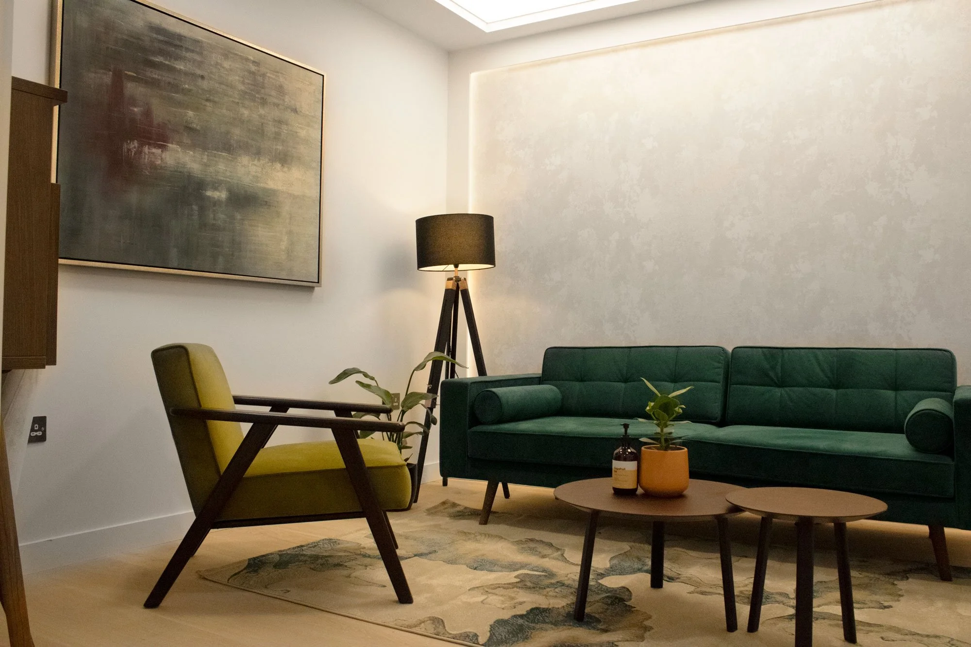 Living room with green sofa, yellow chair, abstract wall art, black floor lamp, and wooden coffee tables with plants and a bottle.