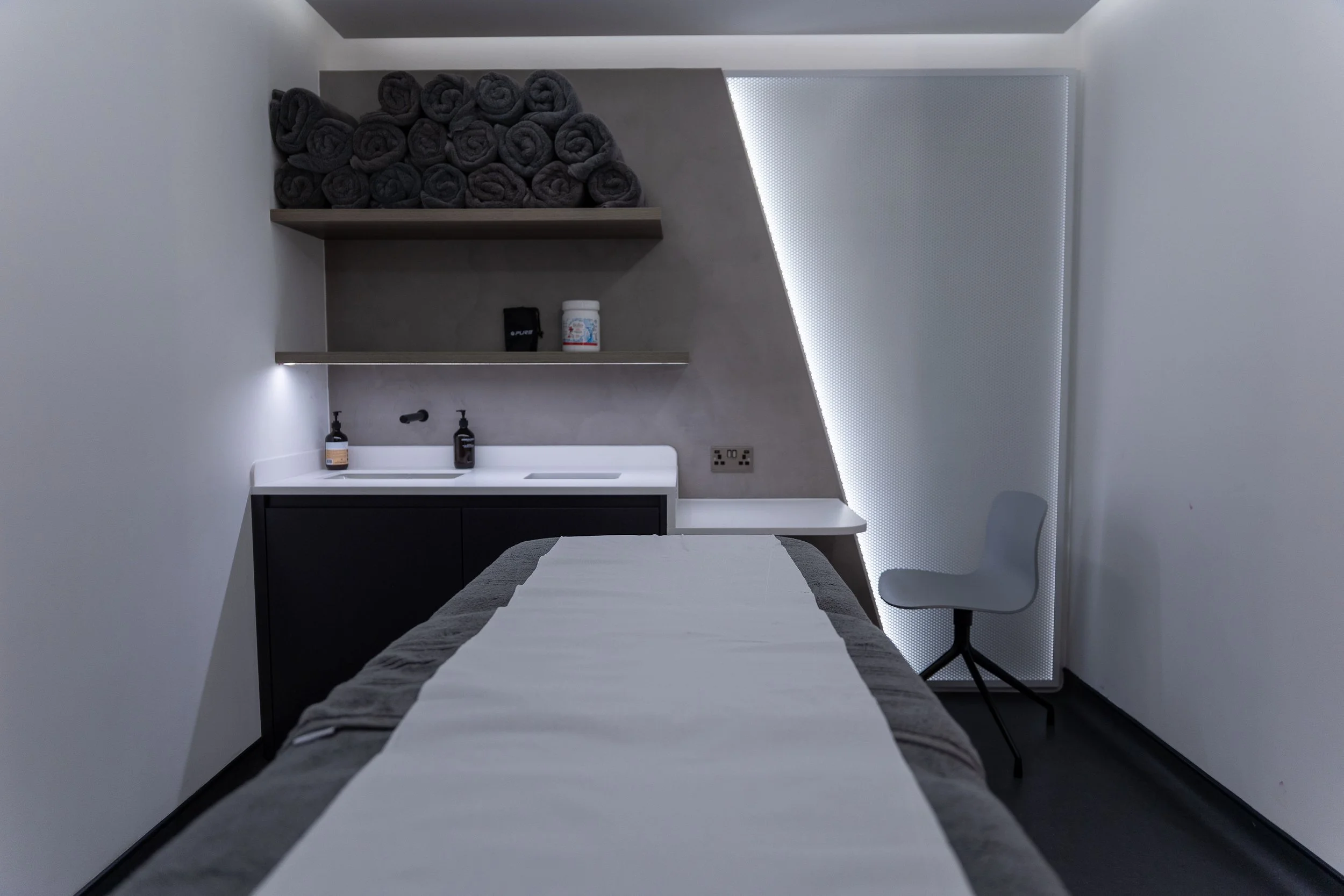 A modern medical examination or treatment room with a massage or treatment bed in the foreground, a cabinet with folded towels on the top shelf, a small sink with soap and sanitizer bottles, a chair, and a sleek, minimalist design with subdued lighti
