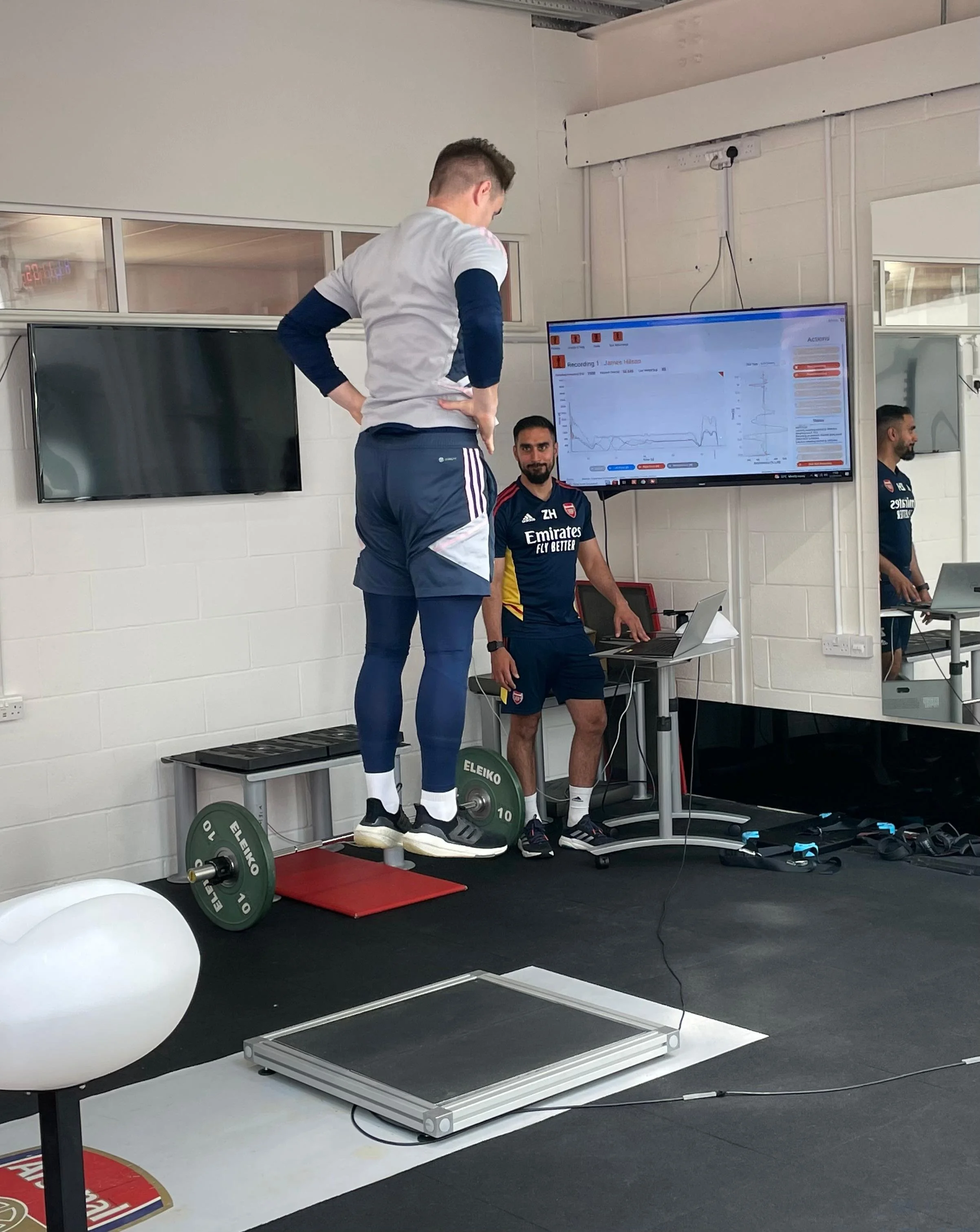 A man is standing on a force plate, undergoing a physical test or assessment, with a technician standing nearby monitoring data on a laptop and a large screen displaying graphs and information. The man is wearing athletic clothing.