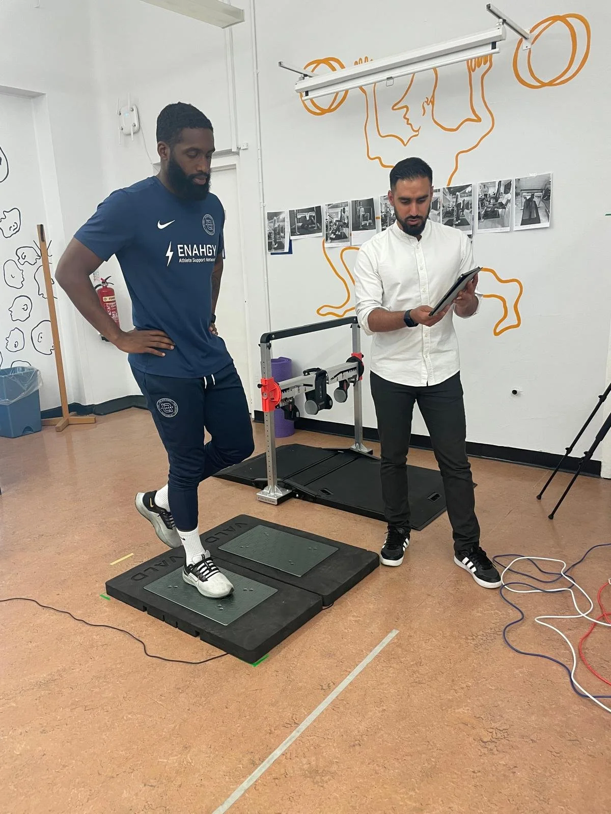 A person in a blue athletic shirt and pants standing on a force plate in a laboratory or sports science facility, with another person in casual clothing holding a tablet and monitoring the person on the force plate.