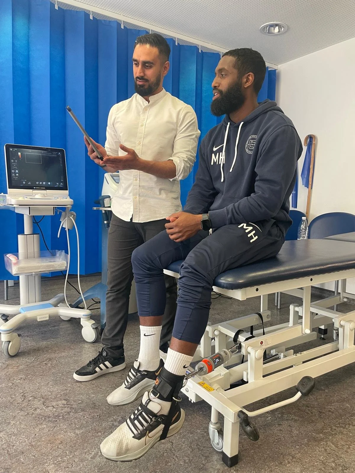 A man in casual athletic clothing is sitting on a medical examination table with his legs secured in a medical device, while another man in a white shirt stands next to him, looking at a tablet. The room has medical equipment and a blue curtain in the background.