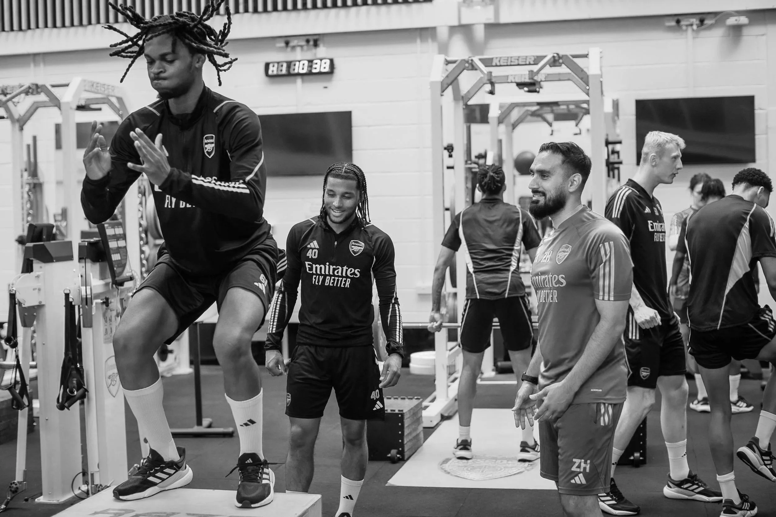A group of soccer players and trainers in a gym, with one player on a step exercise, others smiling and watching, and workout equipment in the background.