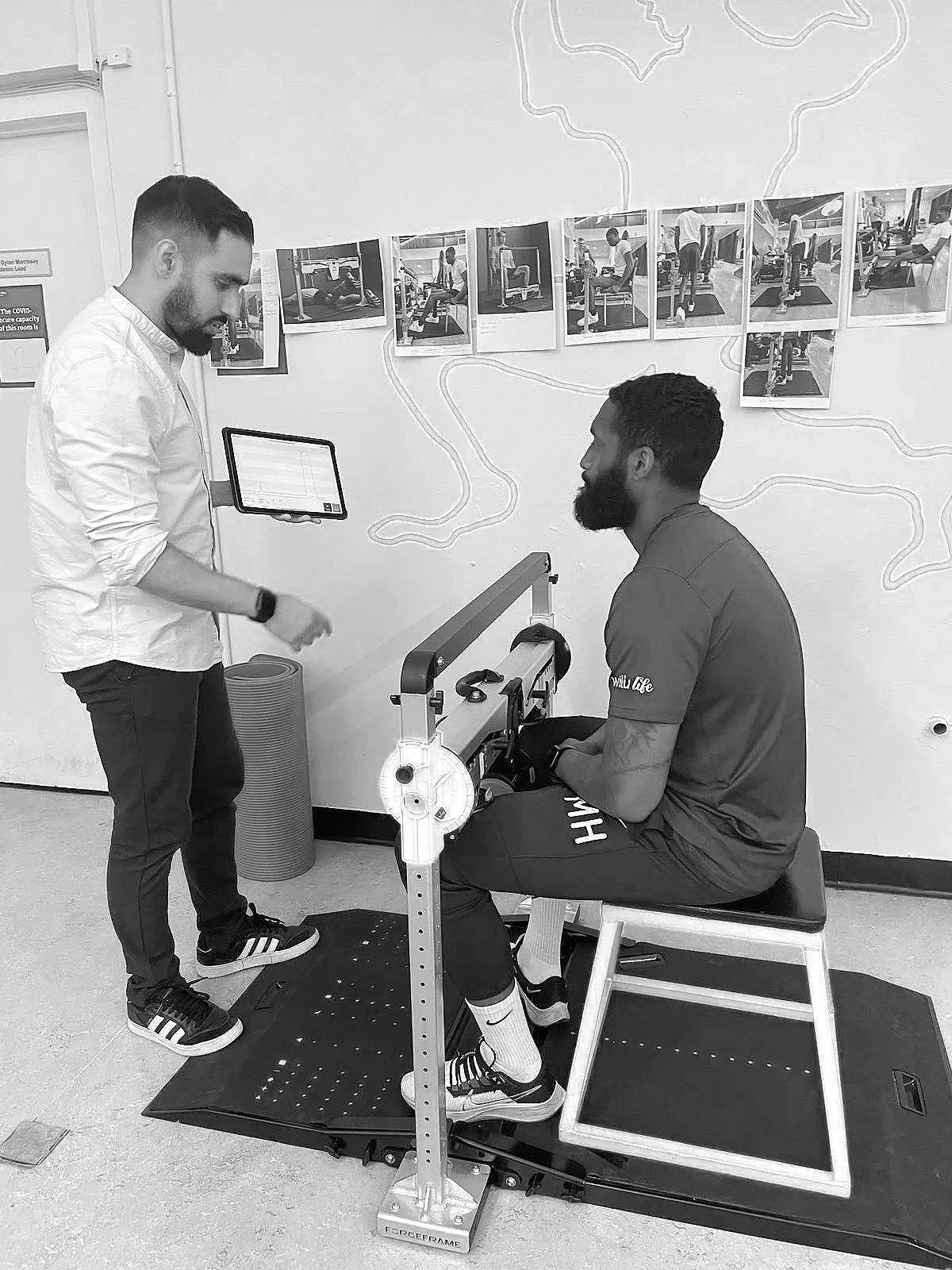 A man sitting on a chair with a leg brace being examined by a therapist using a gait analysis machine, in a clinical setting.