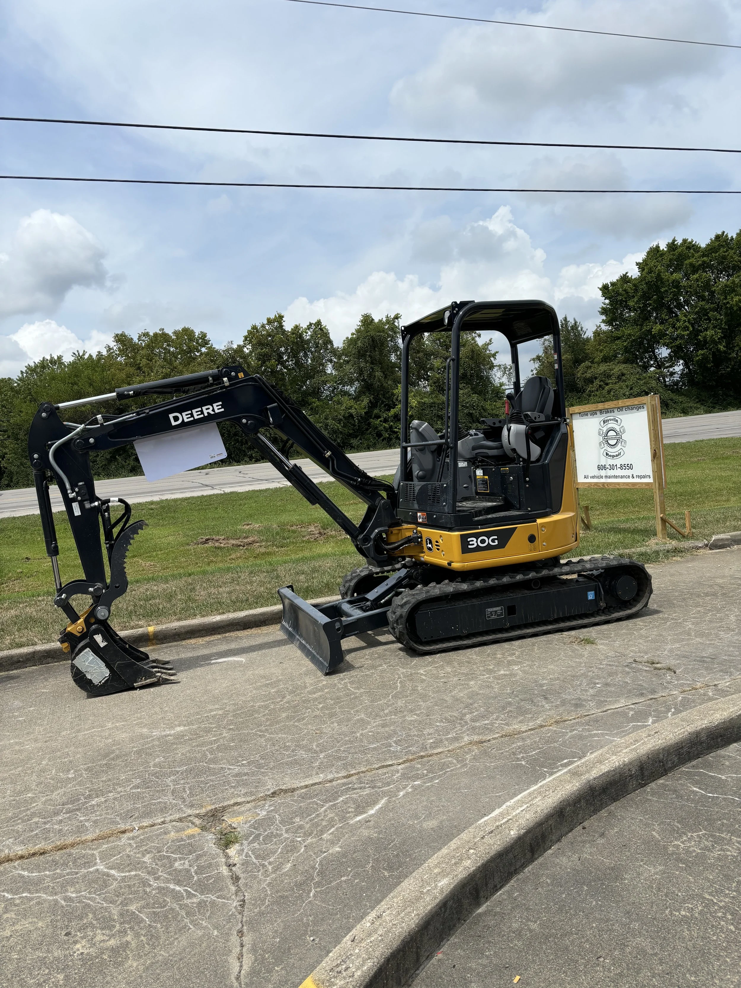Compact excavator parked on concrete, with a sign displaying business information in the background, and trees under a cloudy sky.