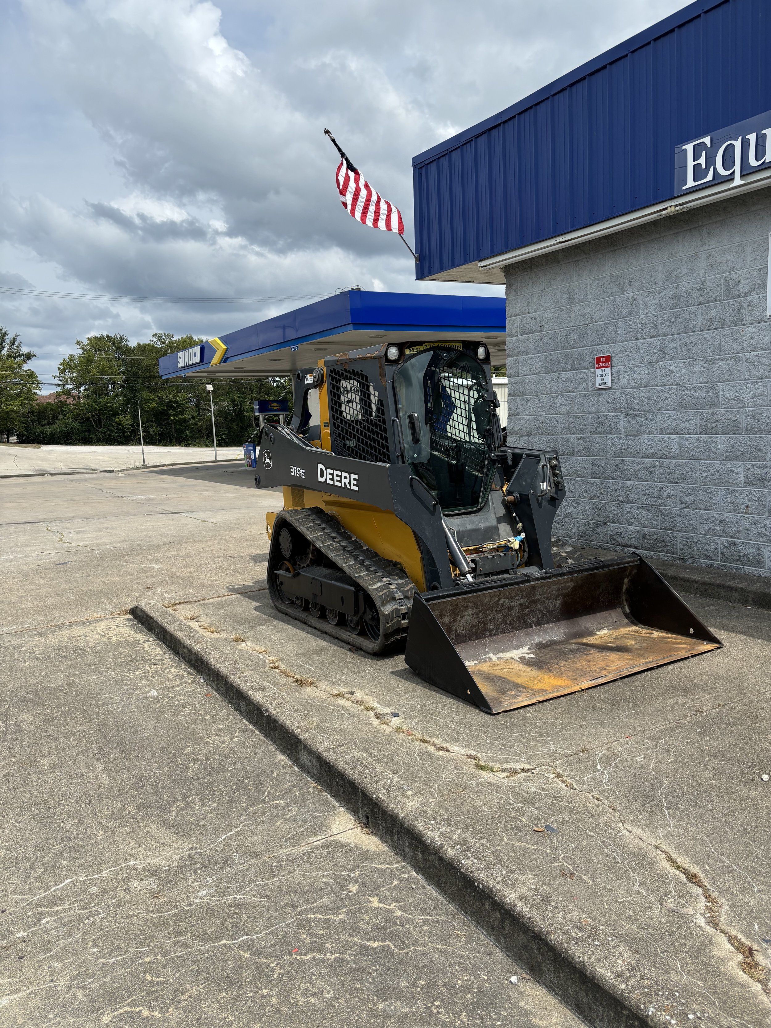 A skid steer loader parked in front of a blue canopy labeled Sunoco, with an American flag flying above.
