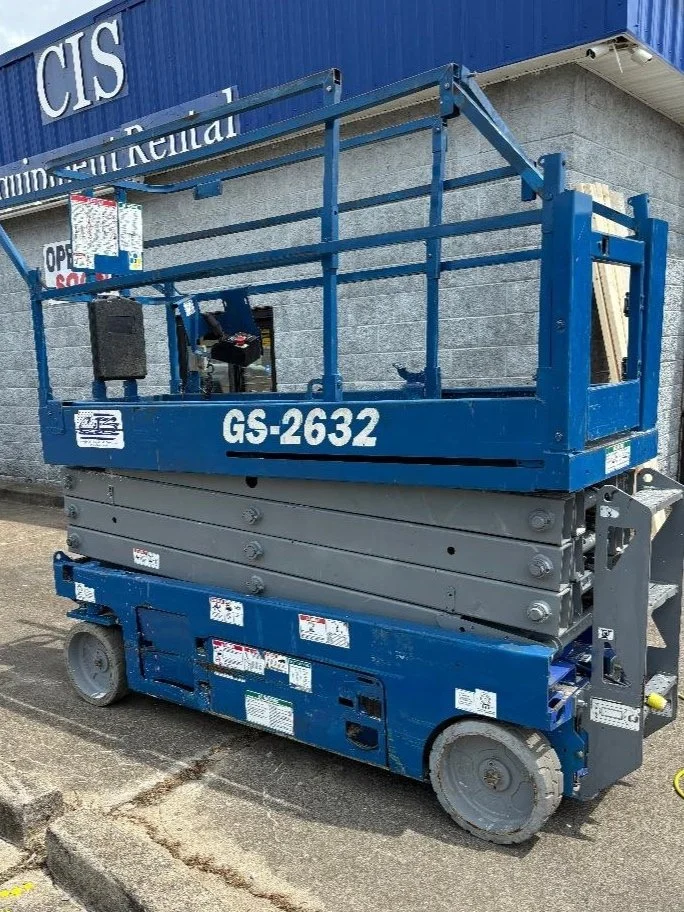 Blue scissor lift labeled GS-2632 parked outside a building with a sign reading 'Equipment Rental.'