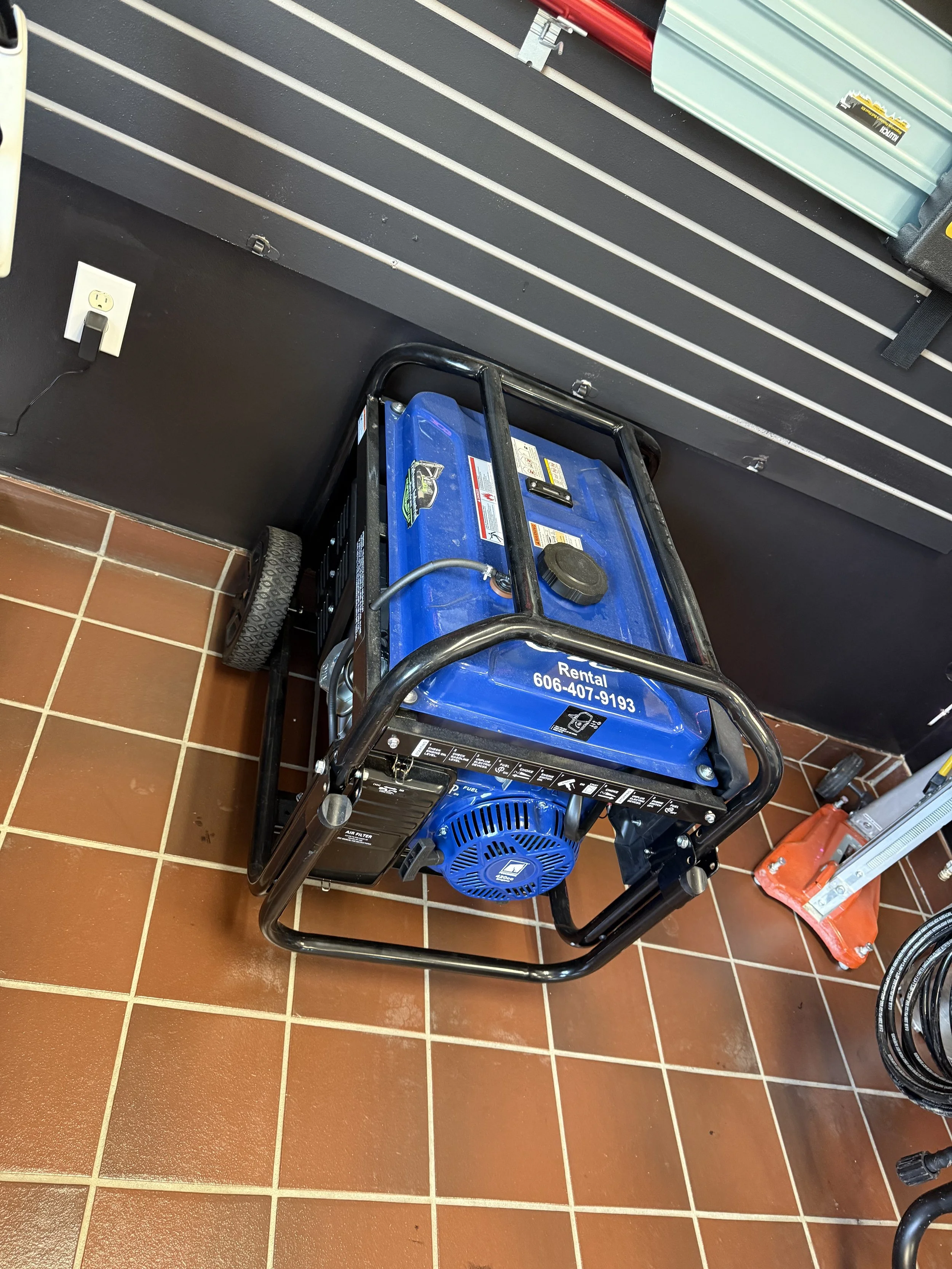 Portable generator with a blue housing and black frame on a tiled floor, positioned near a wall with electrical outlets and other equipment.