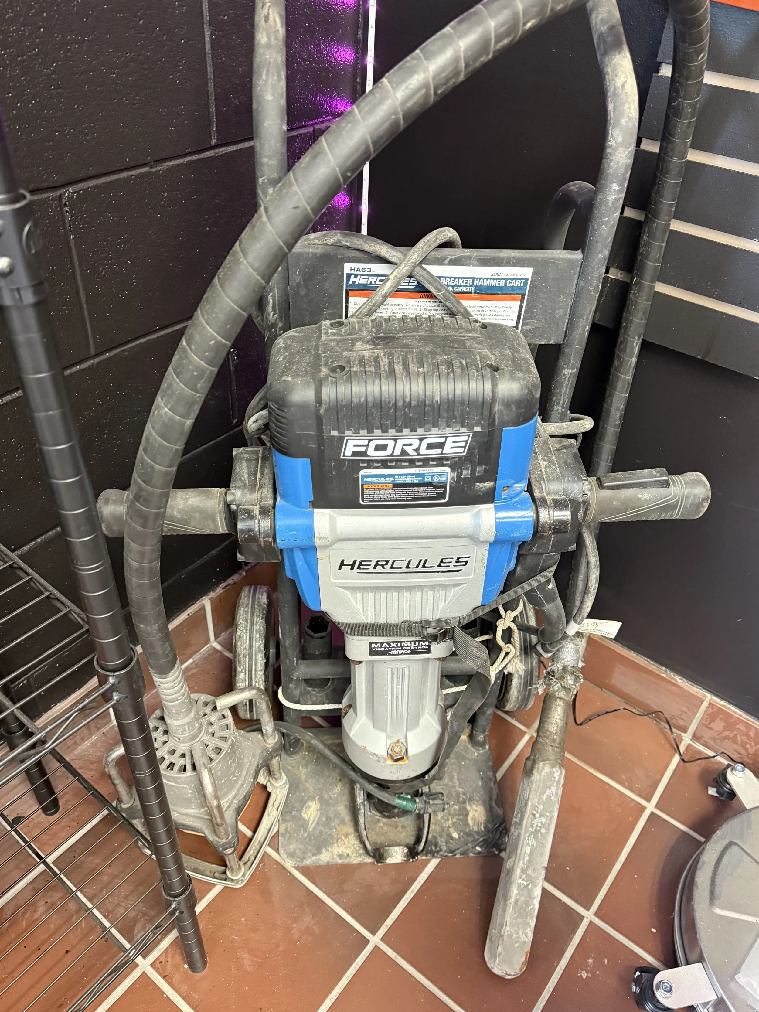 A Hercules heavy-duty demolition breaker hammer secured on a cart, with visible handles and attachments, positioned on a tiled floor against a black wall.