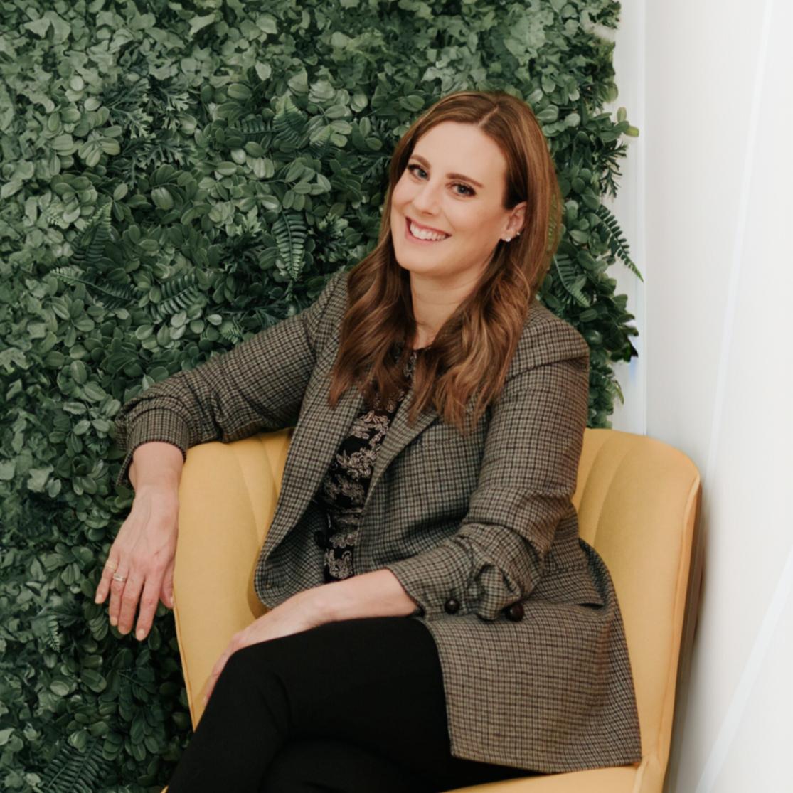 A woman with long, wavy brown hair smiling while sitting on a light yellow chair with a green leafy wall behind her, wearing a brown plaid blazer and black pants.