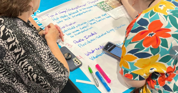 Two people sitting at a table with notes, markers, and a smartphone, engaged in discussion or collaborative work.