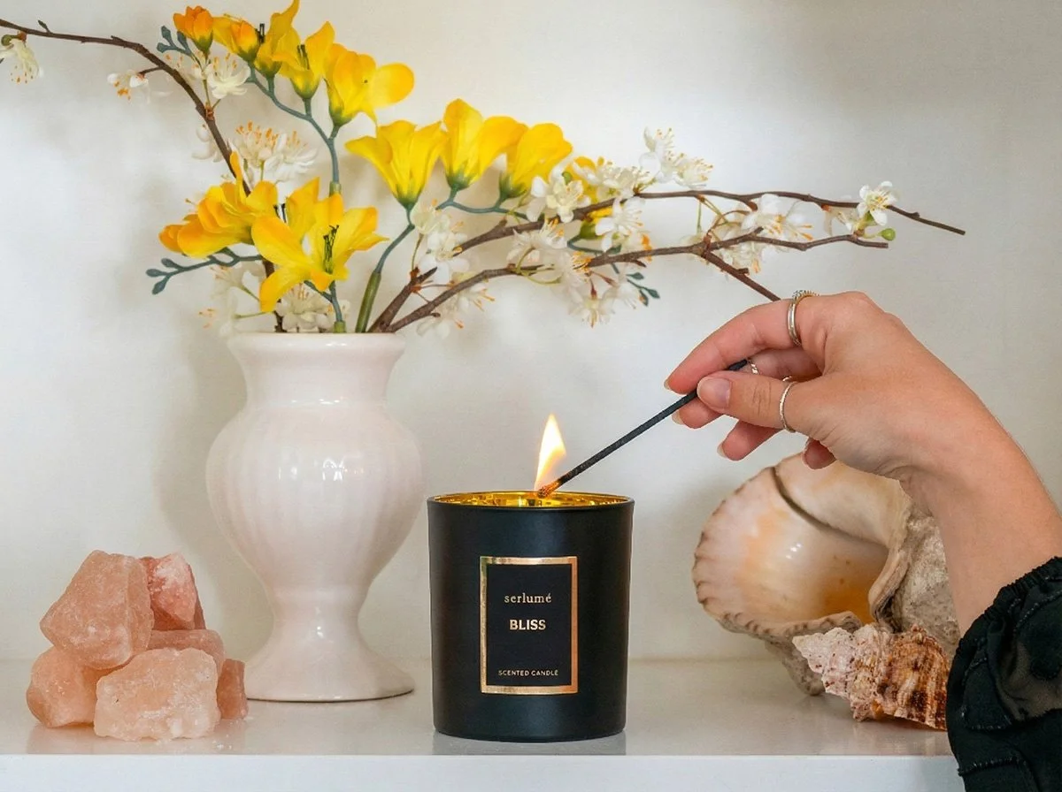 A hand lighting a black scented candle labeled 'Bliss' with a matchstick, with yellow and white flowers in a white vase and seashells and pink salt rocks on a white surface.