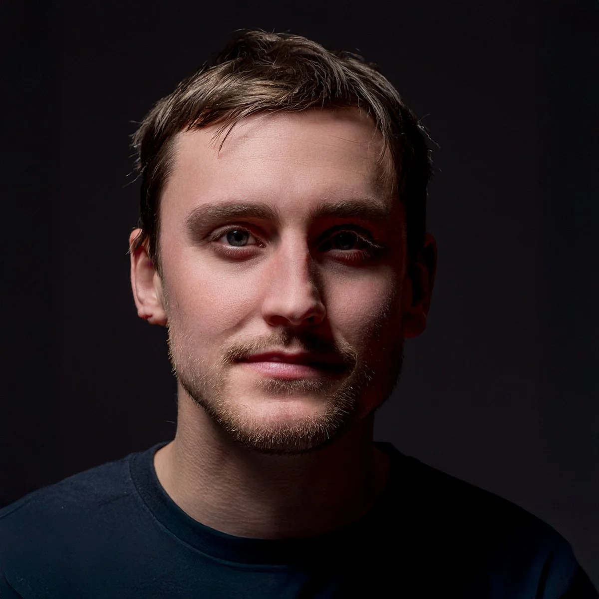 Close-up portrait of a young man with short brown hair, light skin, and blue eyes, wearing a dark shirt against a dark background.