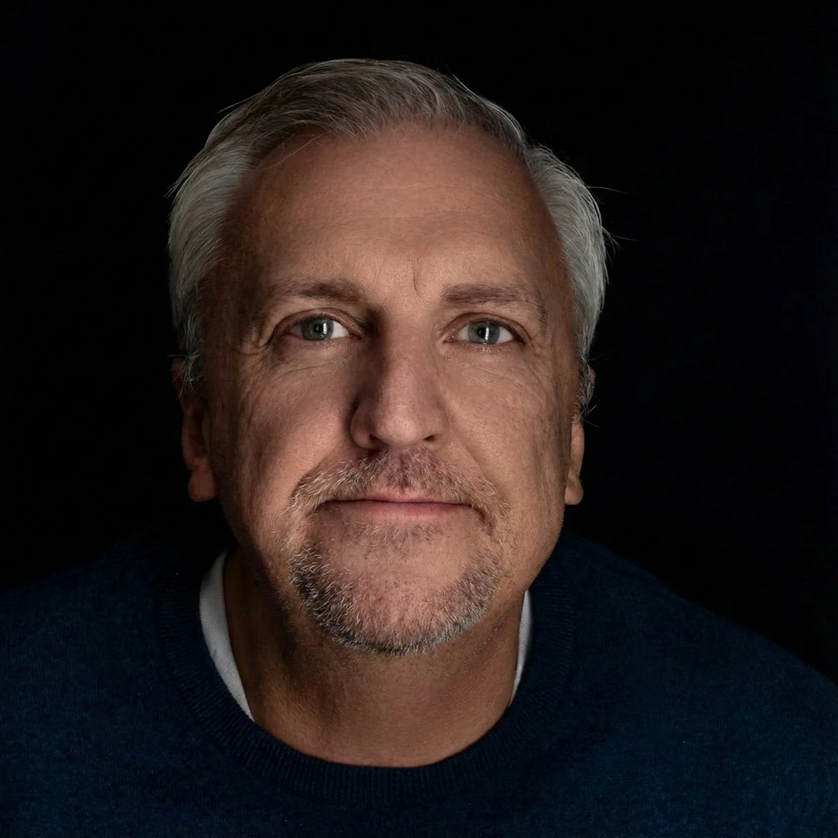 Close-up portrait of a middle-aged man with light skin, gray hair, and a beard, against a black background.