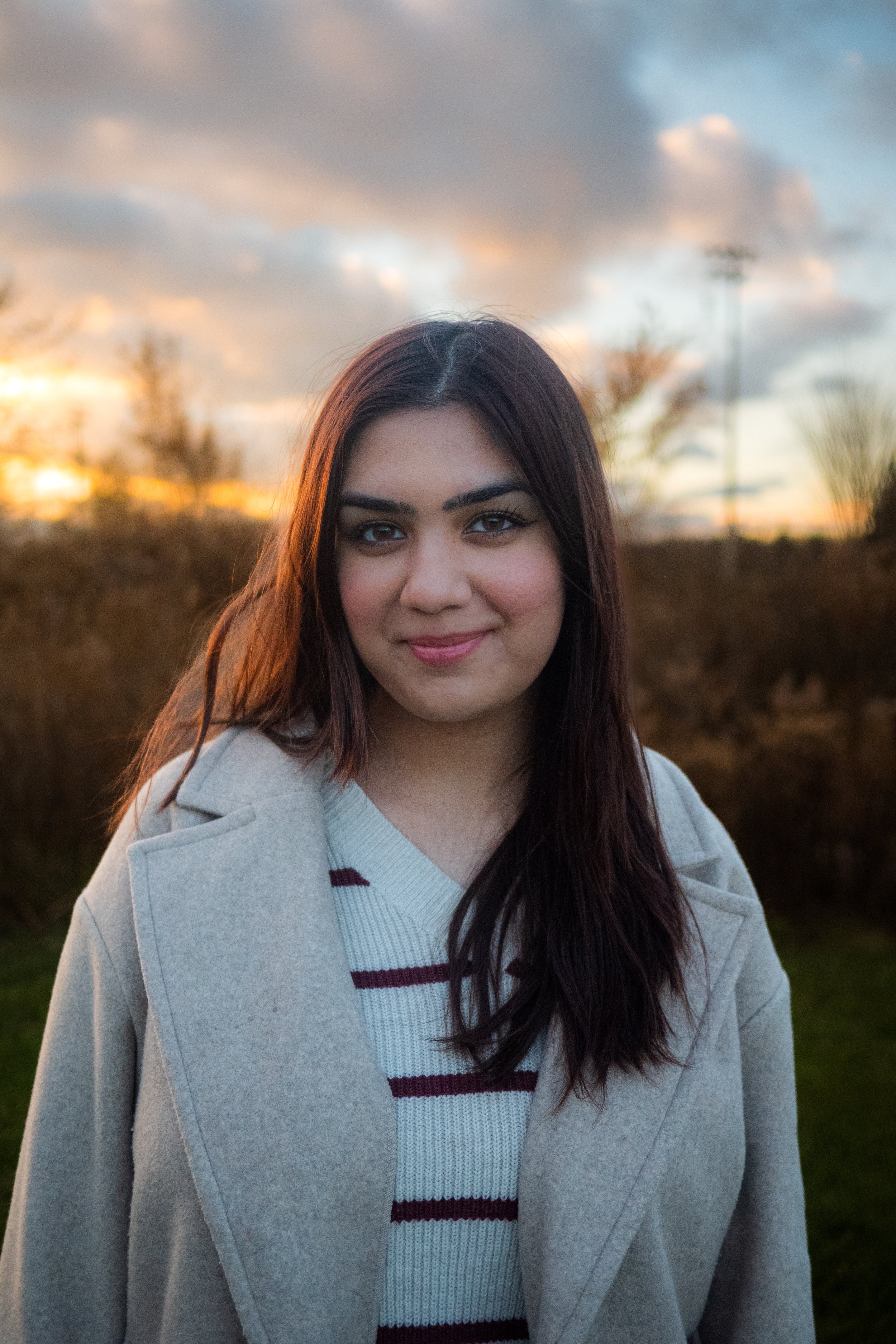 A young woman with dark hair and a light-colored coat standing outdoors during sunset, with a partly cloudy sky in the background.