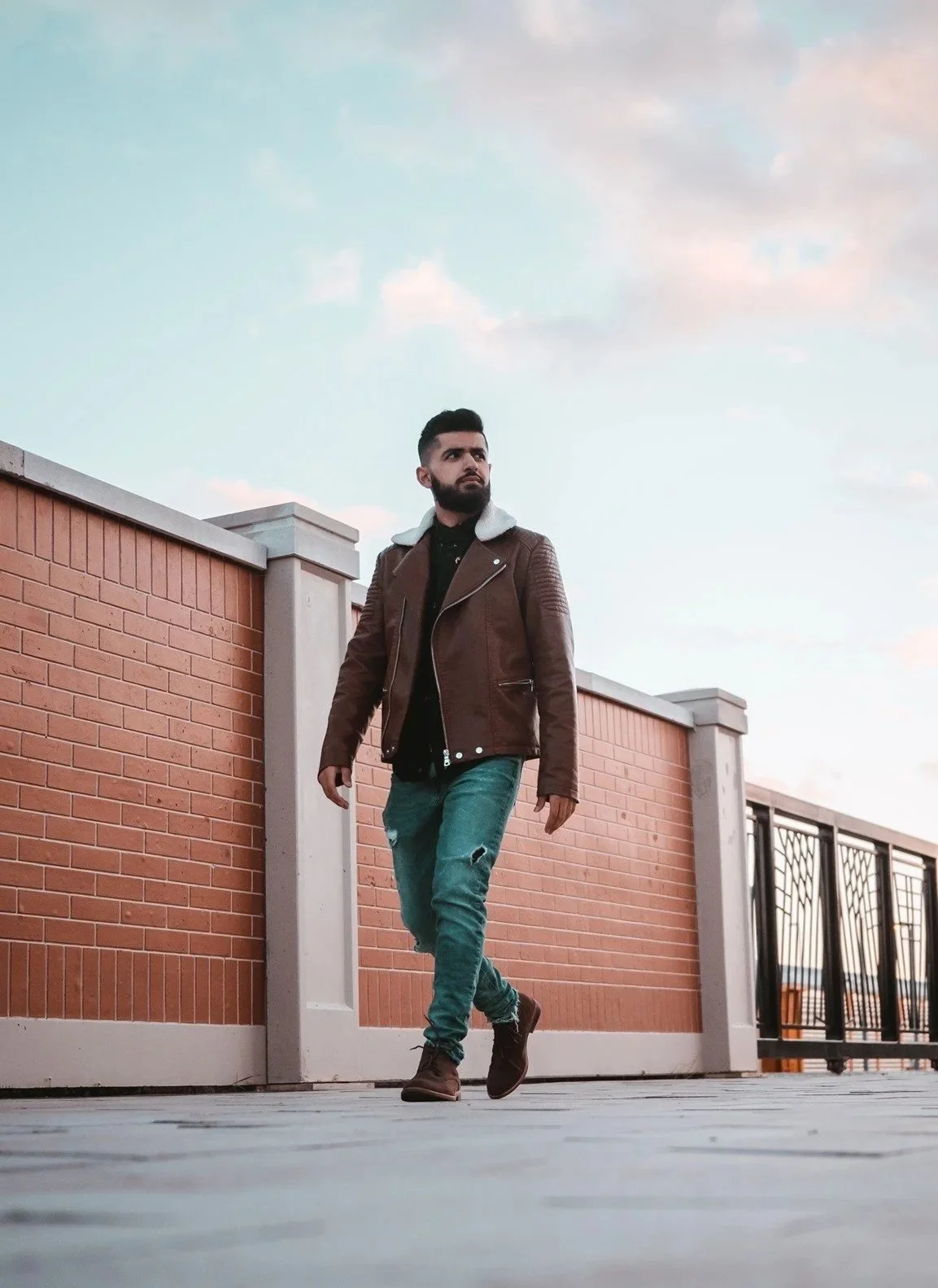 A man with a beard walking on a bridge with brick walls and railings during sunset, wearing a brown leather jacket, black shirt, distressed jeans, and brown shoes.