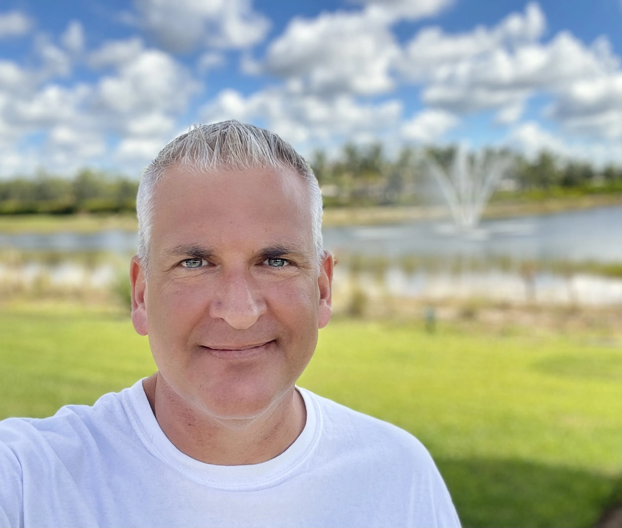 A man with short blonde hair and blue eyes smiling outdoors, with a pond, a fountain, and a partly cloudy sky in the background.