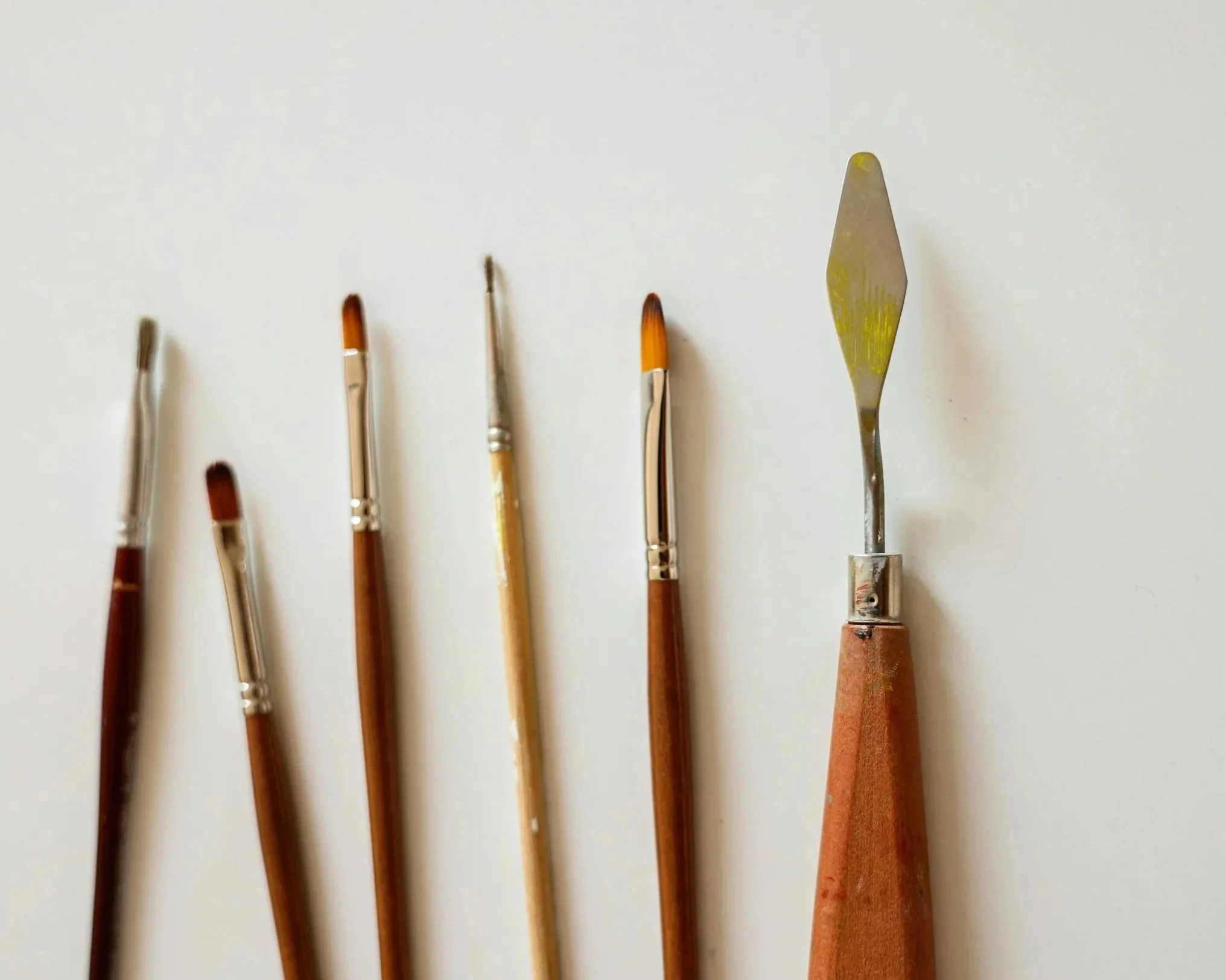 Vijf verfkwasten en een palettemes liggen op een witte achtergrond, allemaal met verfsporen.