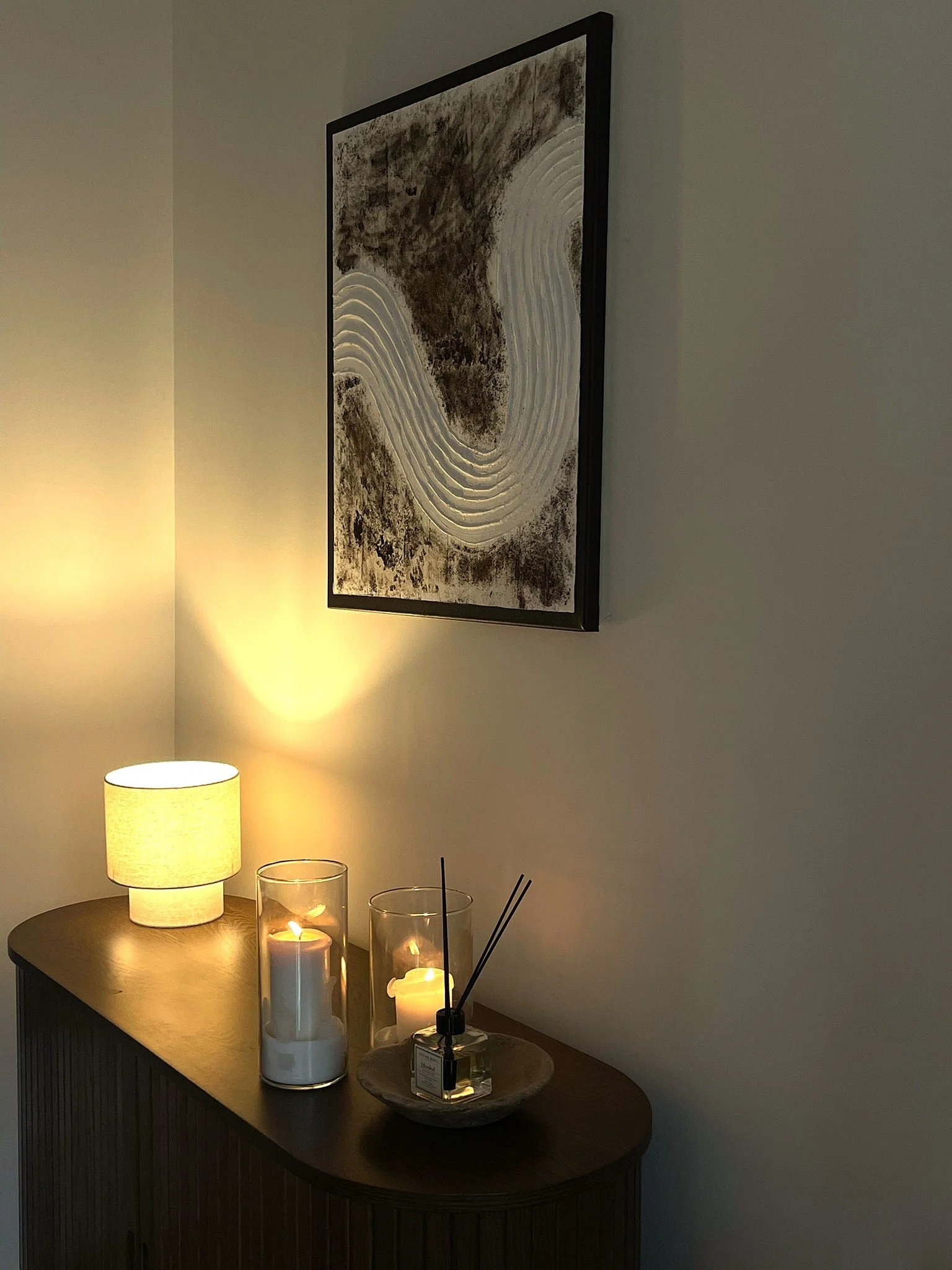 Een houten dressoir met drie kaarsen in glazen kandelaars, een geurstokje, een lamp op de muur en een rechthoekig kunstwerk aan de muur met organische vormen.