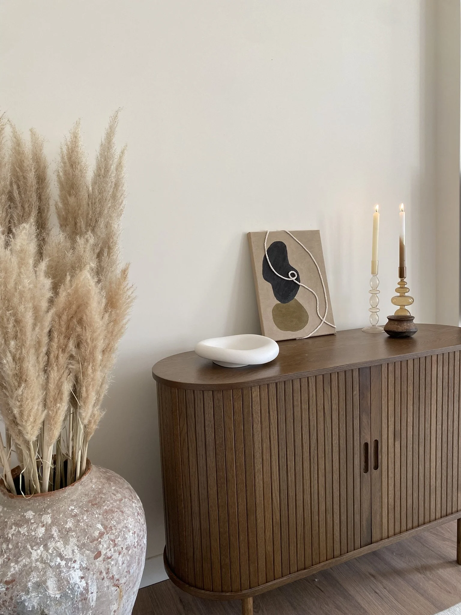 Een houten sideboard met decoratieve voorwerpen, waaronder een abstract kunstwerk, twee brandende kaarsen en een wit schaaltje, naast een grote pot met droge pampasgras.