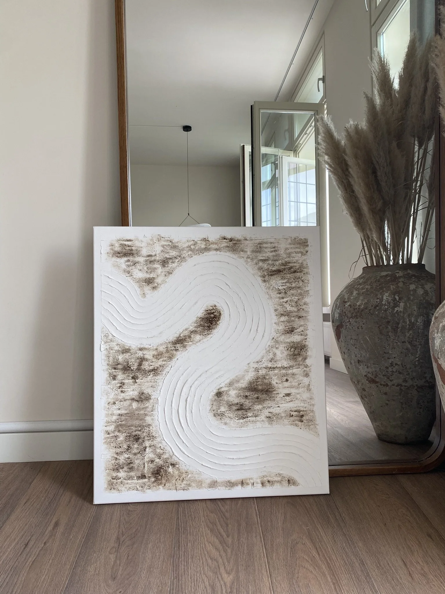 Een wit canvas met een abstract patroon in bruin en wit, gelegen op de vloer voor een muur met spiegel en grote vaas met gedroogde planten.