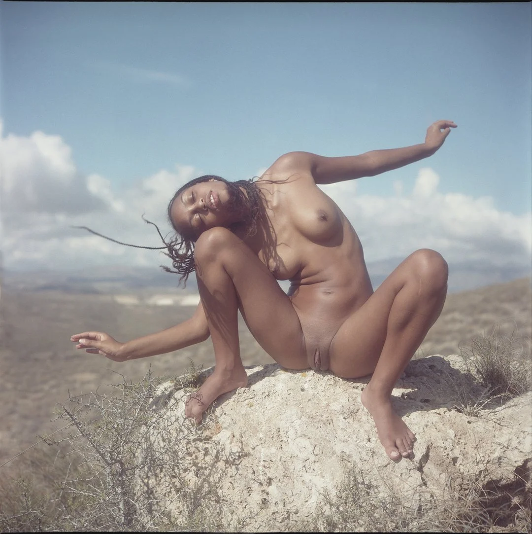 A nude woman with dreadlocks is sitting on a rock in a desert landscape, with her eyes closed and a relaxed expression, under a partly cloudy sky.