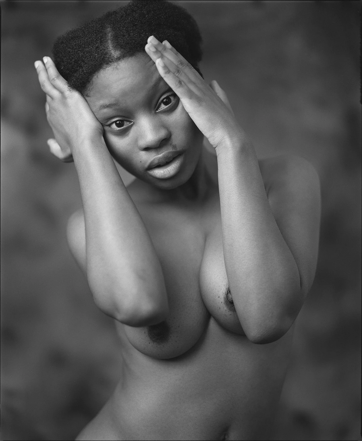 A black and white photograph of a nude woman with short natural hair, holding her head with her hands, with a surprised or contemplative expression.