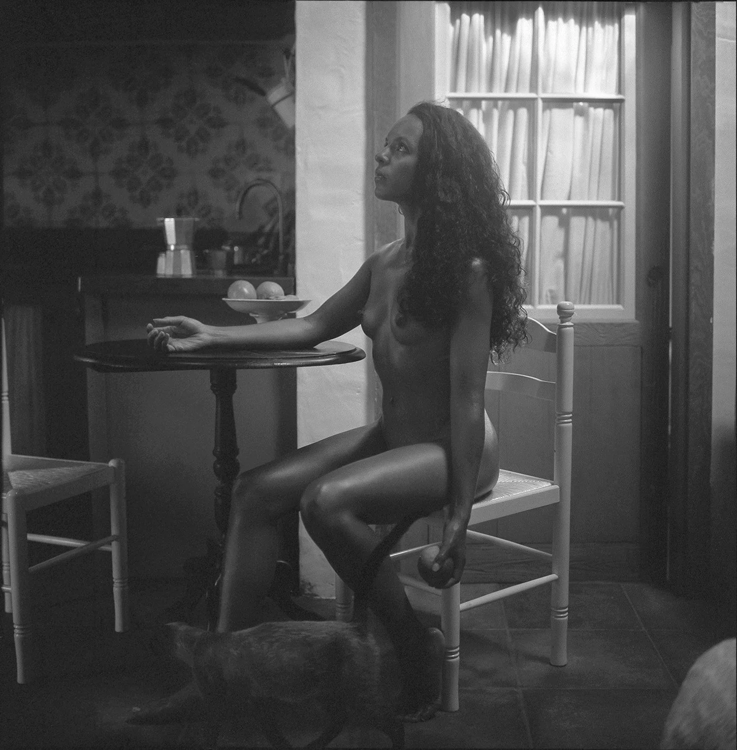 Black and white photograph of a nude woman with long, curly hair seated sideways on a chair in a kitchen, holding a candle in her right hand with a serious expression. There is a dog lying under her feet, a small table with a bowl and two eggs, and a